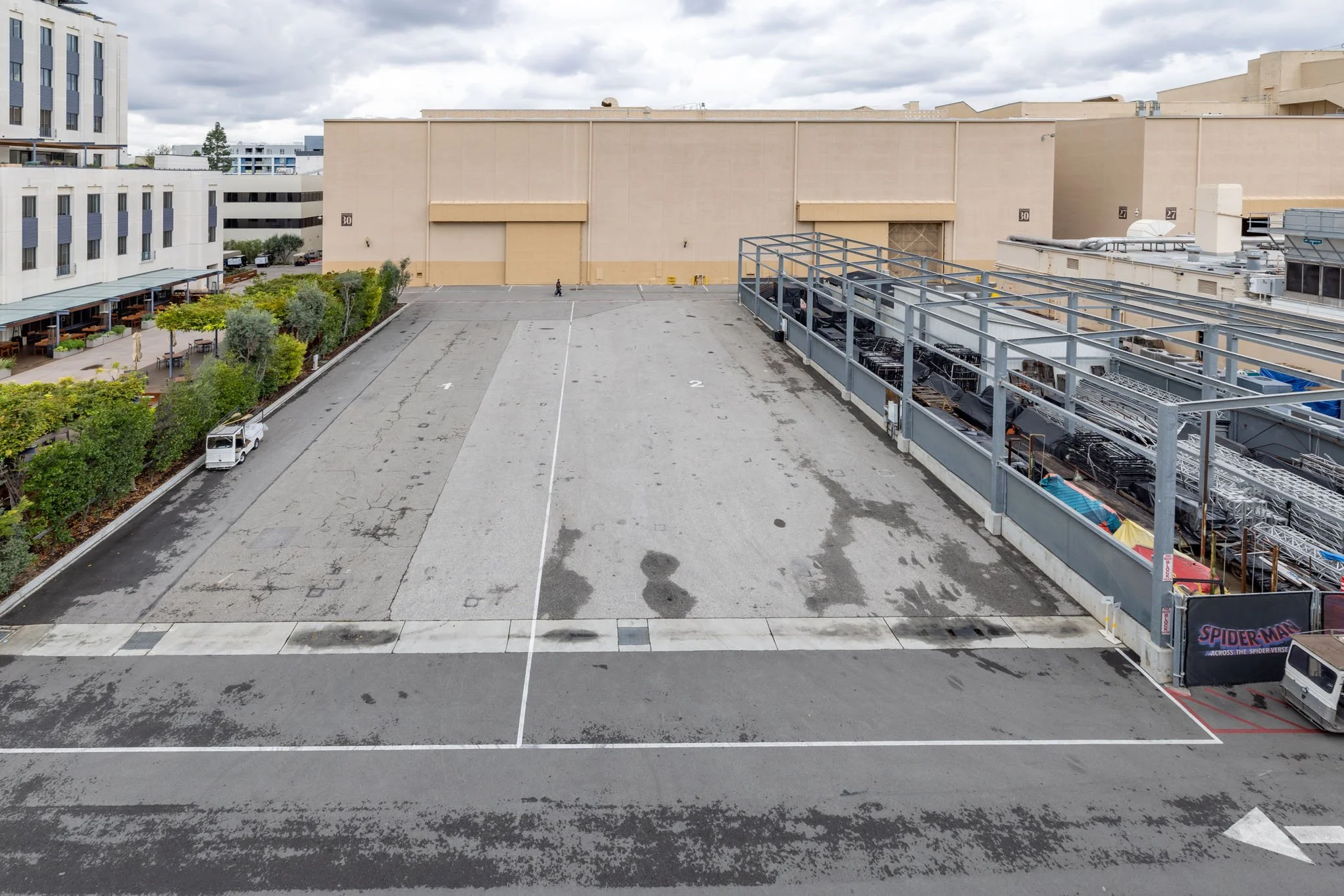 Empty parking lot with few small vehicles, surrounded by trees on the left and a building with a large, beige wall at the back, with some construction materials on the right side.