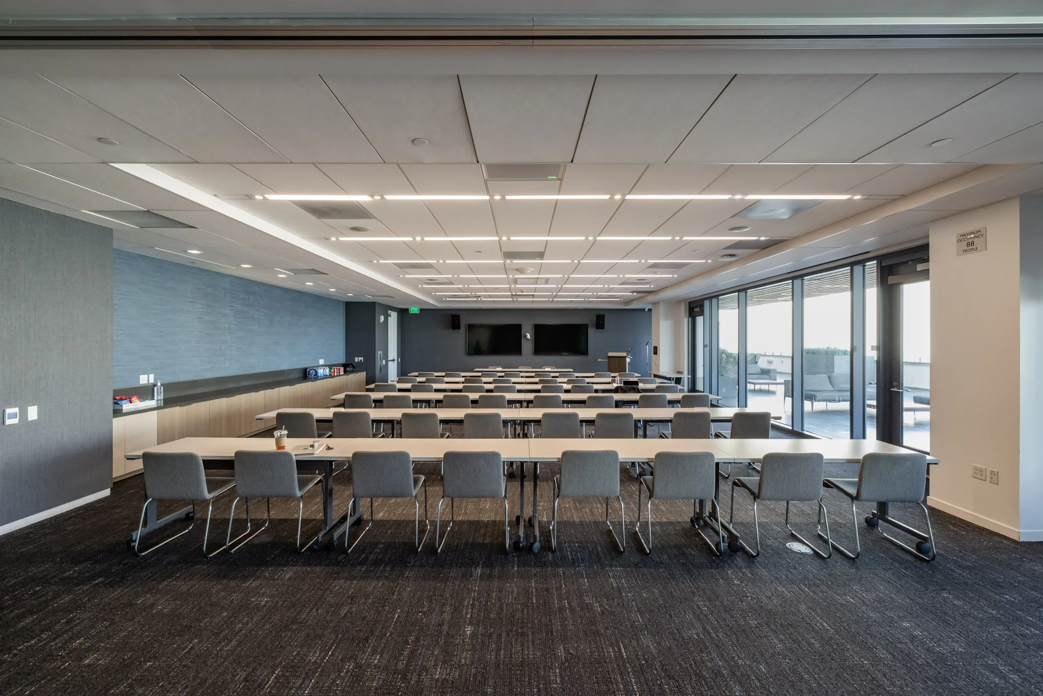 Empty conference room with rows of tables and chairs, two wall-mounted screens at the front, large windows with a balcony outside, and ceiling lights