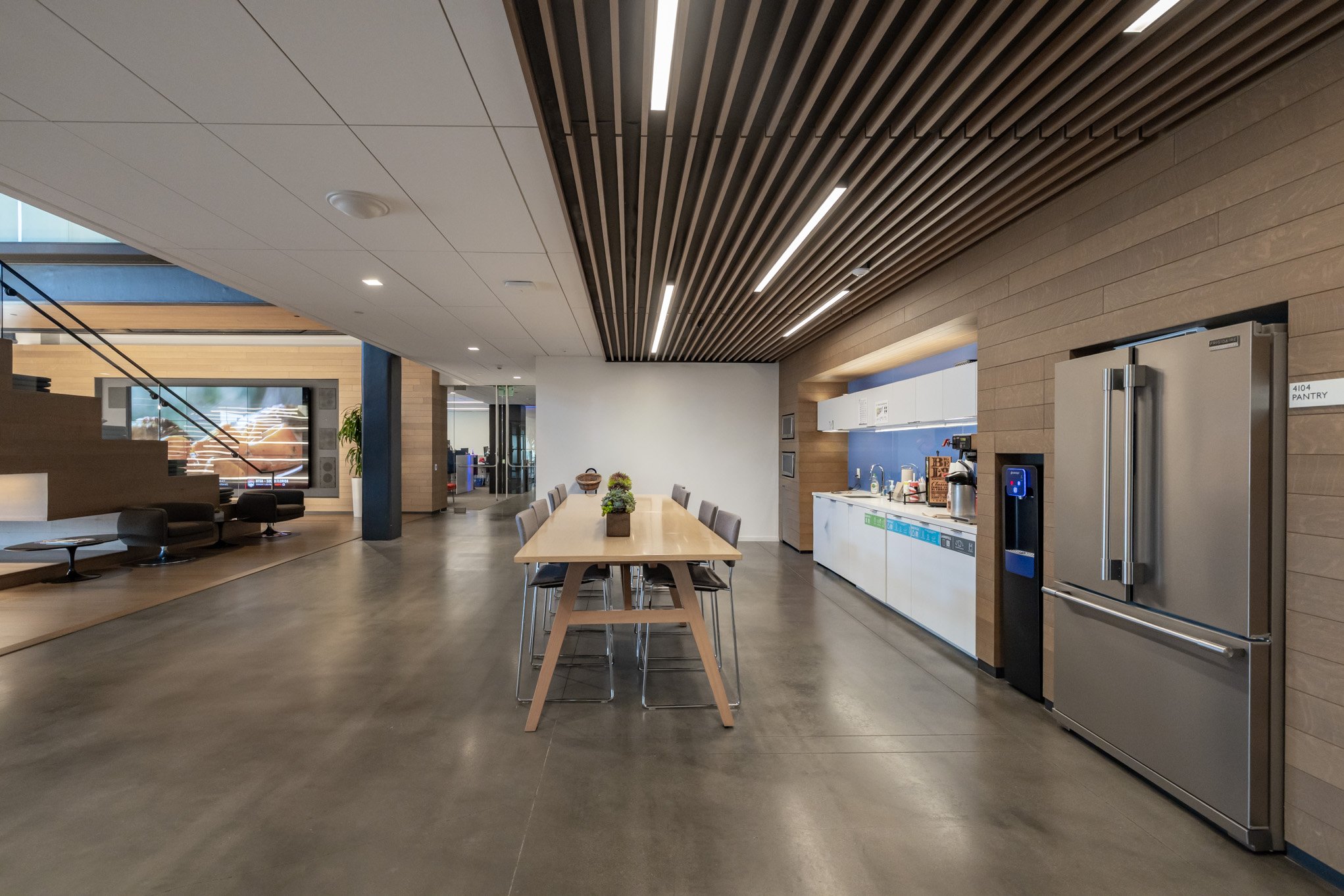 Modern communal kitchen and lounge area with a long dining table, chairs, a refrigerator, microwave, and coffee station, featuring wood accents and built-in lighting.