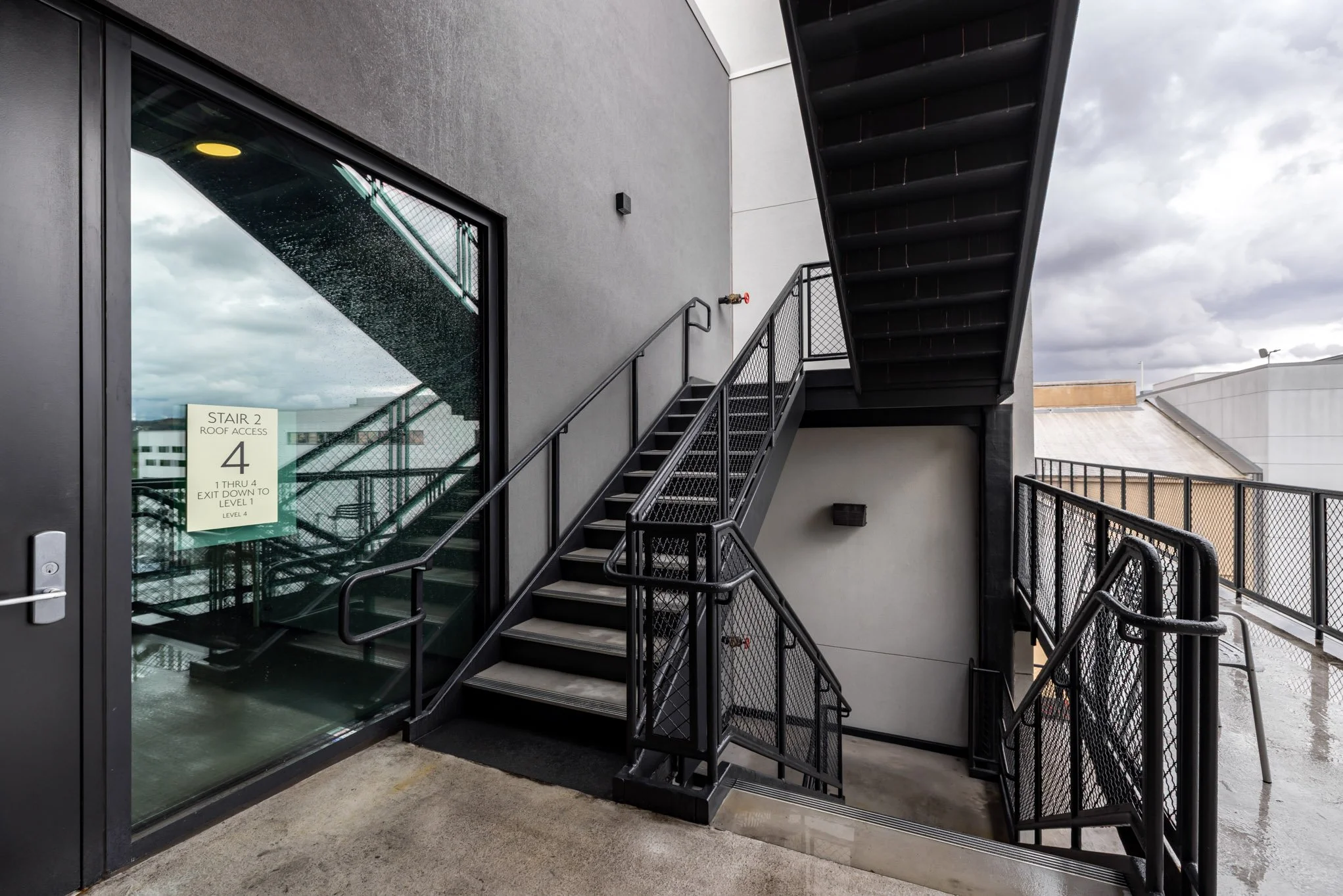 Exterior staircase with black metal railings and stairs leading up to an upper floor, adjacent to a glass door with a sign indicating "Stair 2 Roof Access, 4, 1 through 4 exit down to Level 1," on a rainy day.