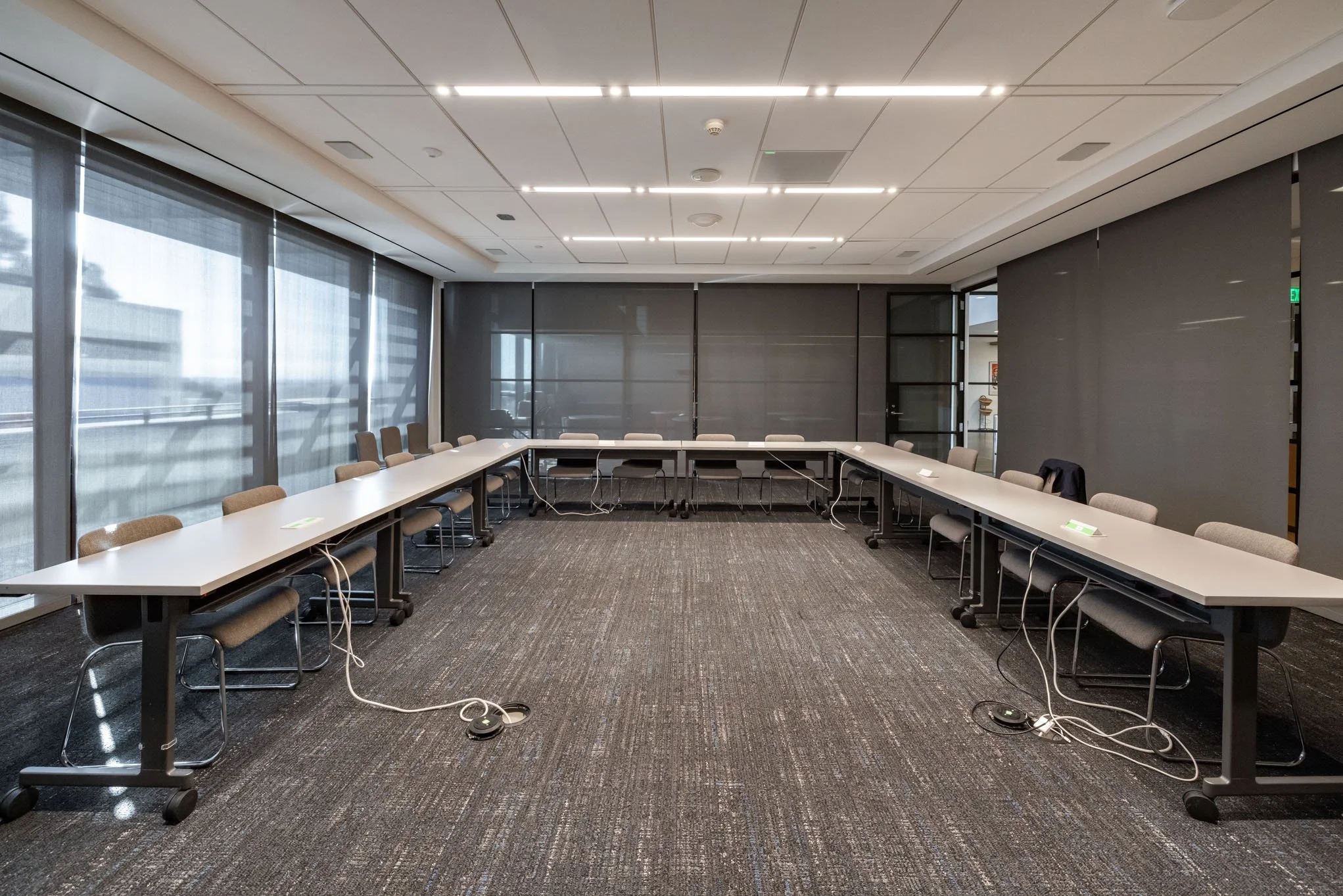 Empty conference room with U-shaped white tables, black chairs, large windows with blinds, and a gray carpet, with visible power cords on the floor.