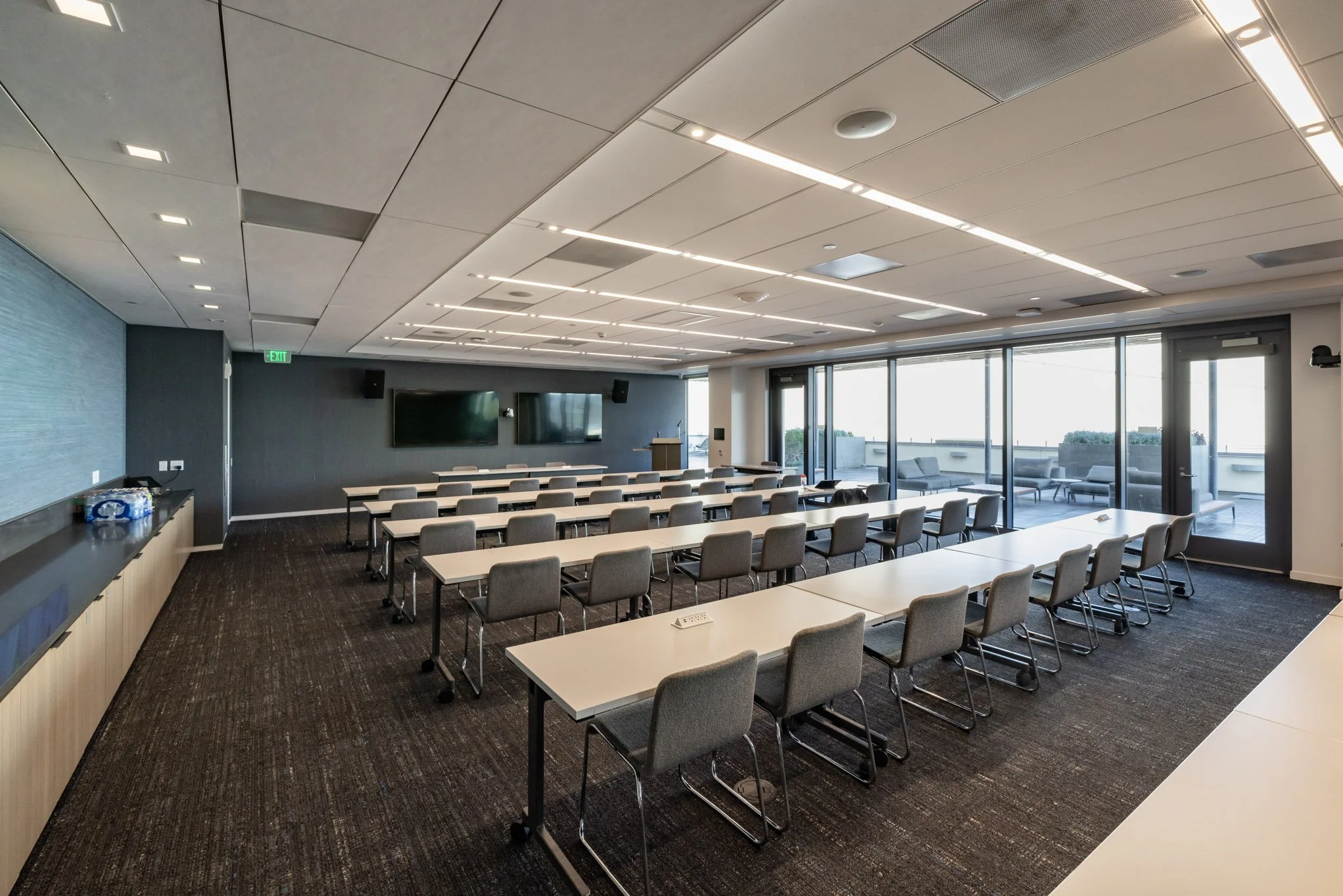 Empty conference room with long desks and chairs, large windows, two TVs at the front, ceiling lights, and a far outdoor terrace.