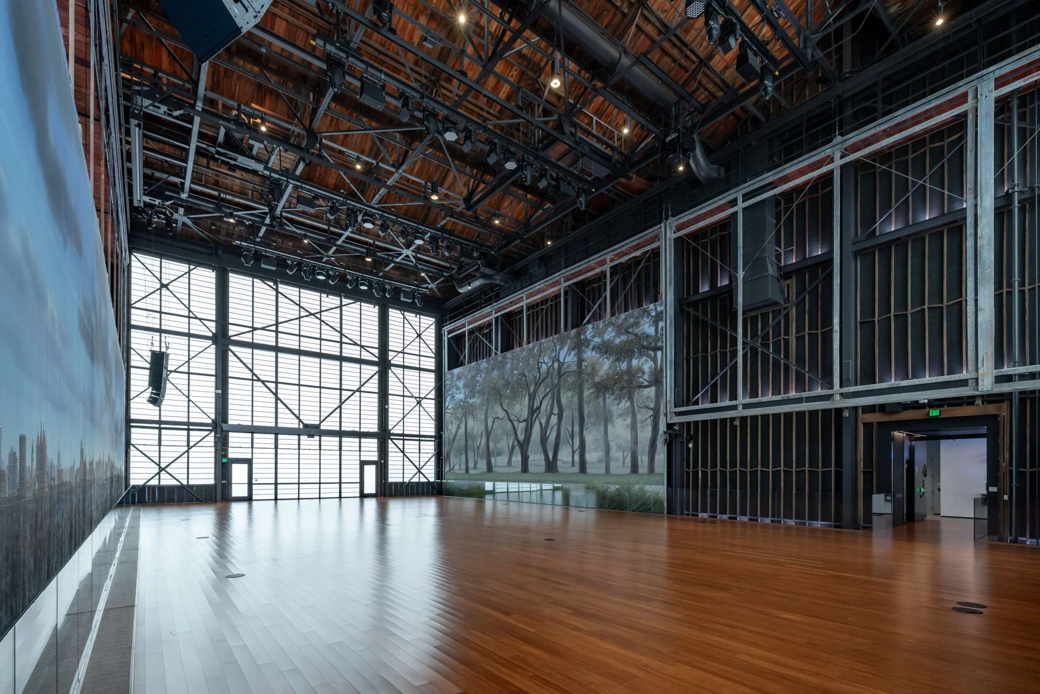 An empty modern indoor gallery space with wooden flooring, a large window wall, a mural of trees, and visible industrial ceiling structure with lighting equipment.