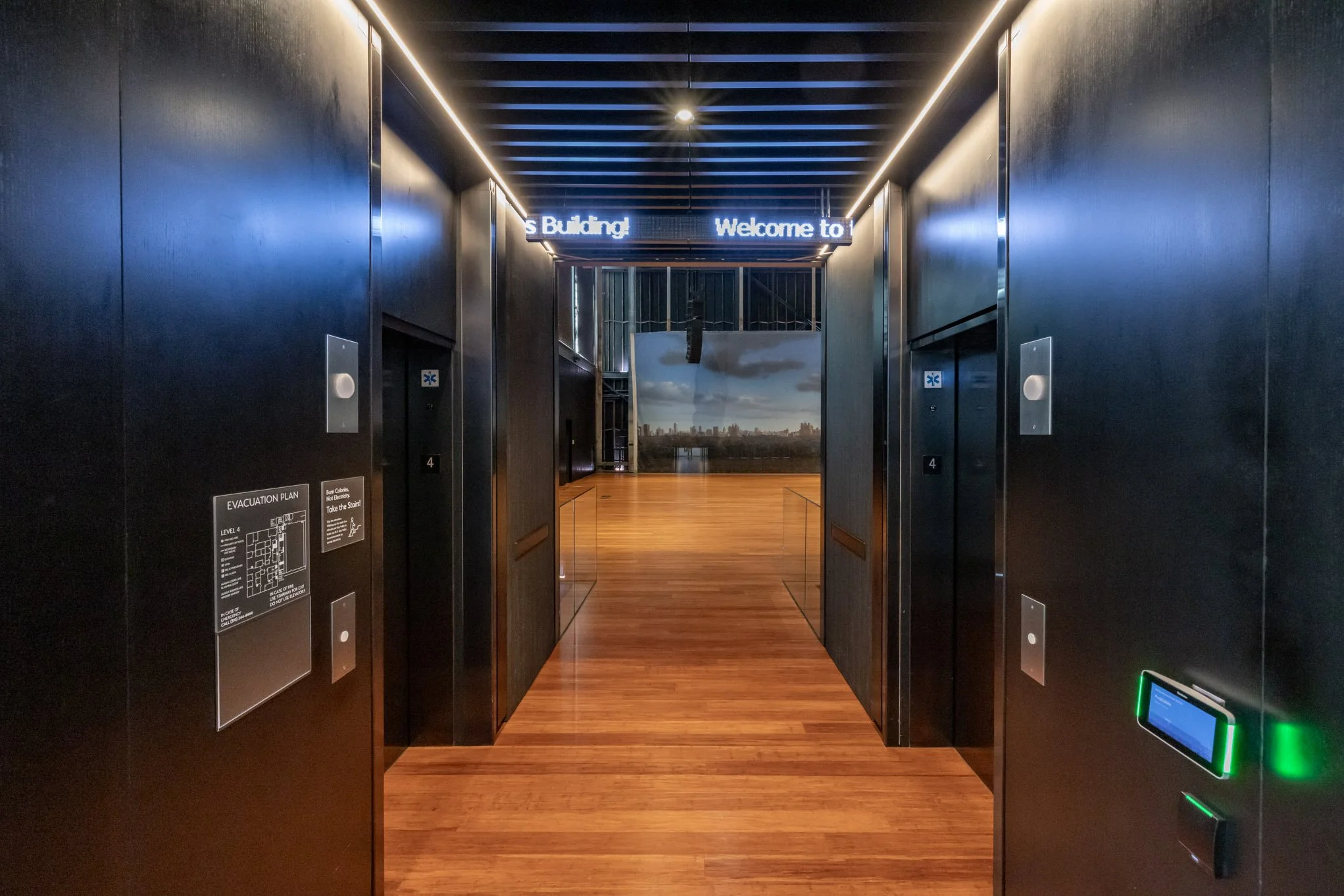 Elevator doors open revealing a hallway with wood flooring and a cityscape window in the background, with a sign that reads 'Welcome to the Building' above the entrance.