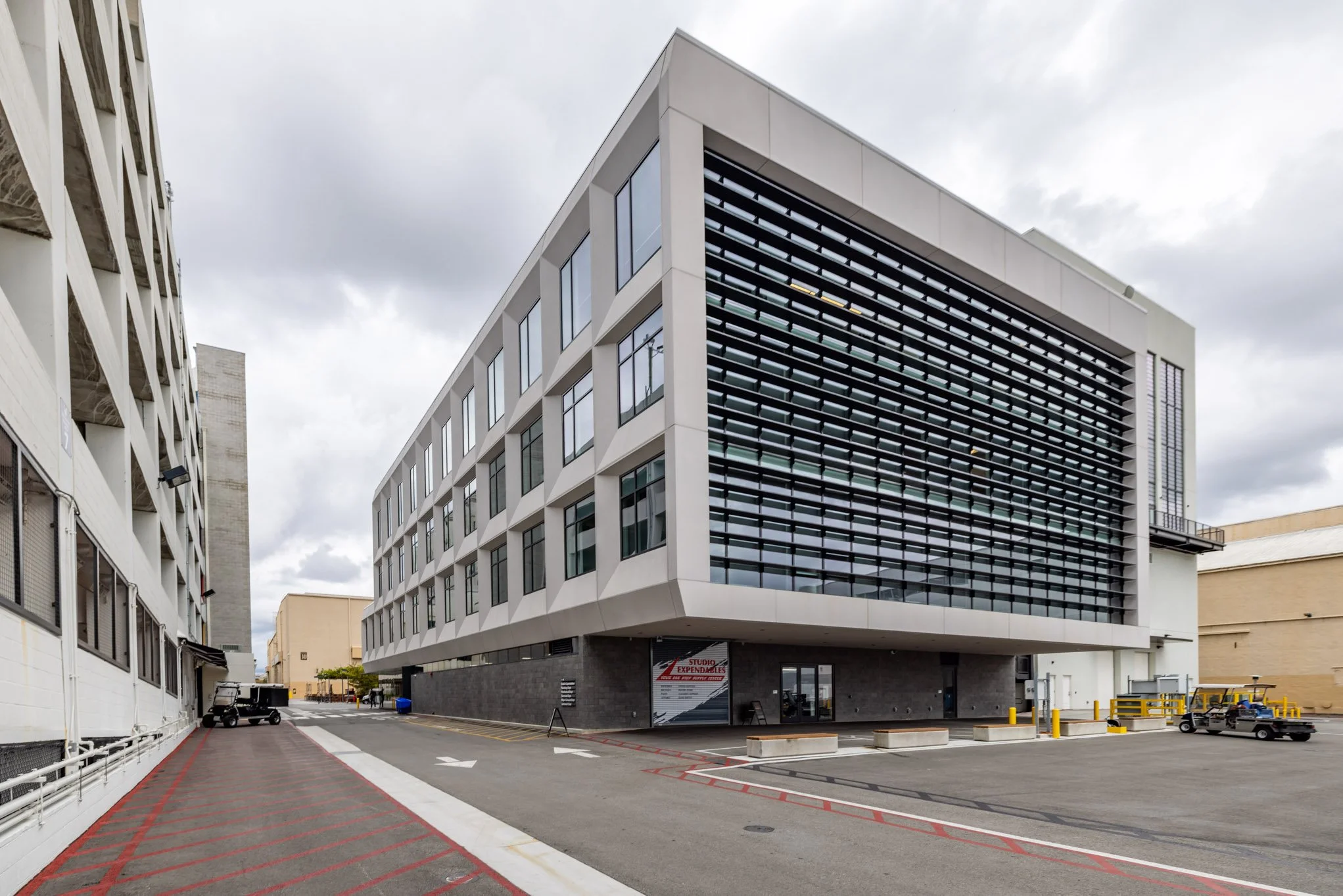 Modern multi-story office building with a distinctive black grille facade, large windows, and a parking area with golf carts outside under a cloudy sky.