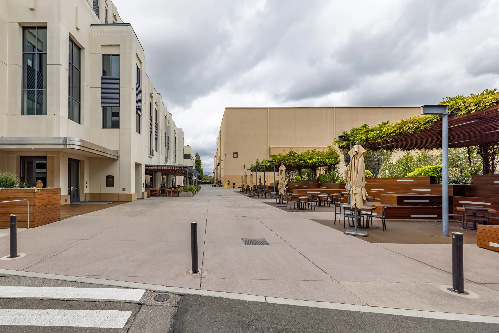 An outdoor urban scene showing a wide sidewalk or plaza between modern buildings with light-colored facades, small plants in planters, benches, tables, and umbrellas on the right side, overcast sky.