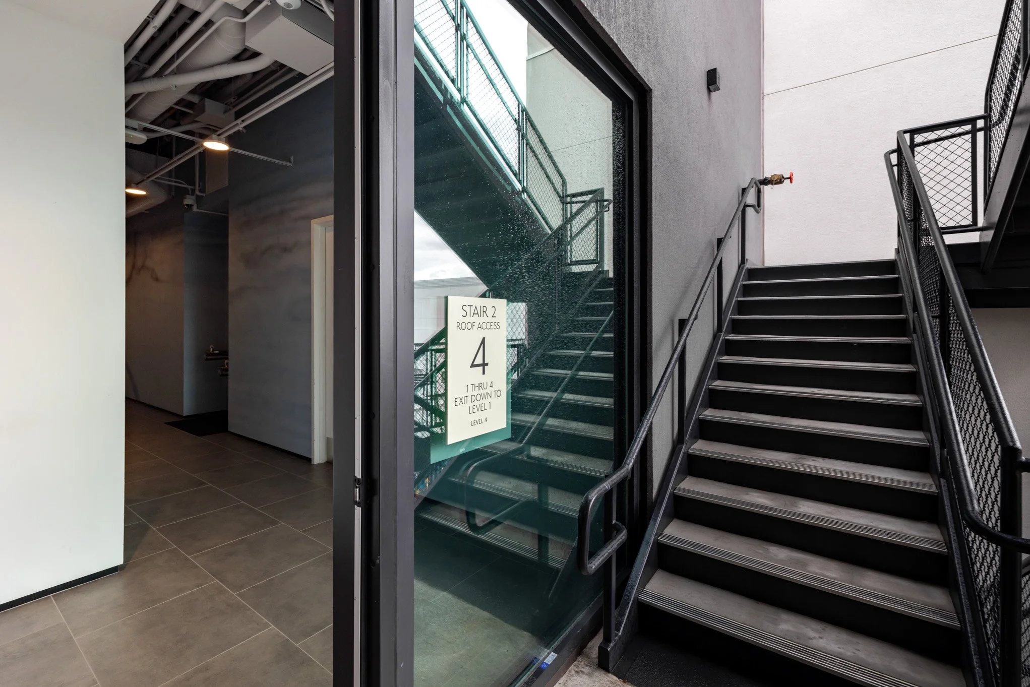 Indoor staircase with a sign on the glass door indicating 'Stair 2 Roof Access, 4, 1 through 4 Exit Down to Level 1, Level 4' with metal railings and a partly covered exterior staircase visible through the glass.