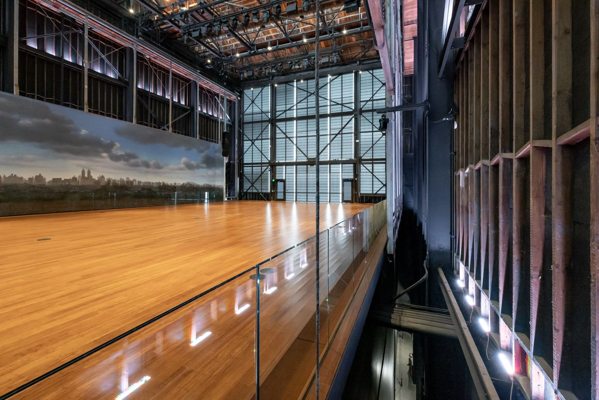 An empty stage with a wooden floor, located inside a theater or performance venue with black metal framing and a cityscape backdrop on one side.