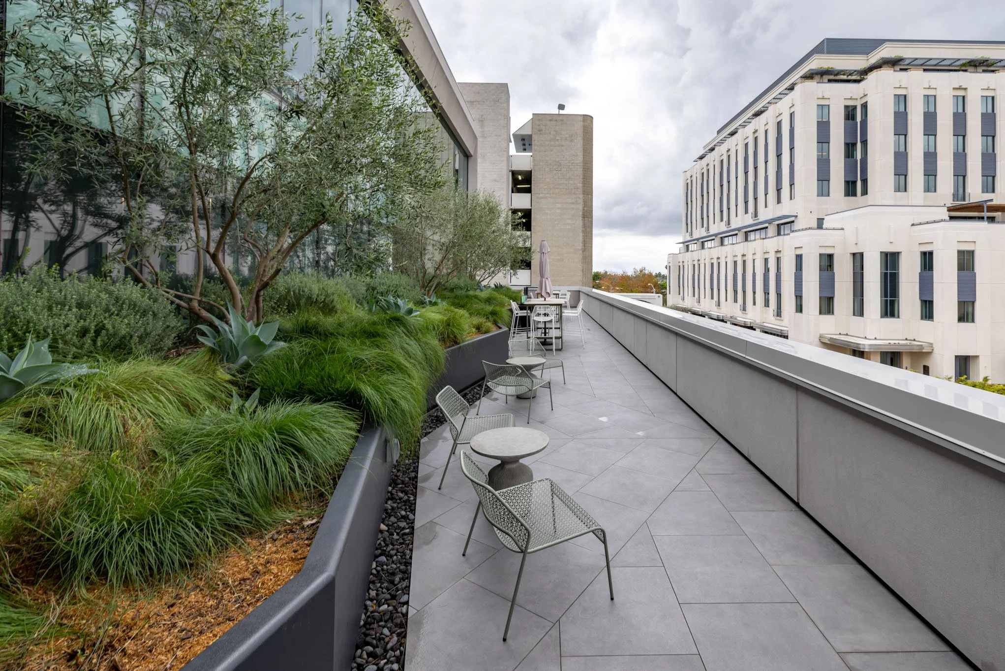 Empty outdoor balcony with green plants, trees, and dry grass in large planters on the left, and metal chairs and tables along with a closed umbrella on the right, with modern buildings in the background under cloudy sky.