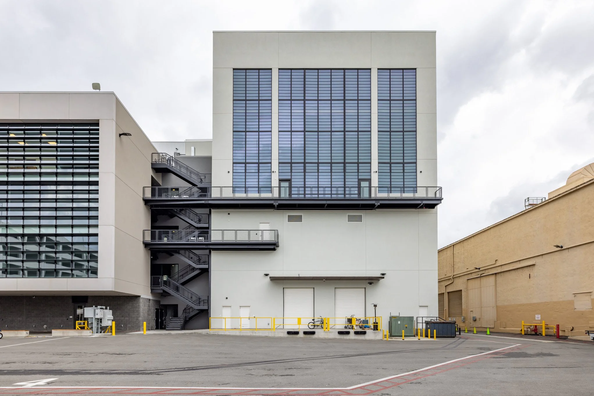 Back of a large industrial or commercial building with a loading dock area, a bicycle, and safety barriers in the foreground, and a cloudy sky overhead.