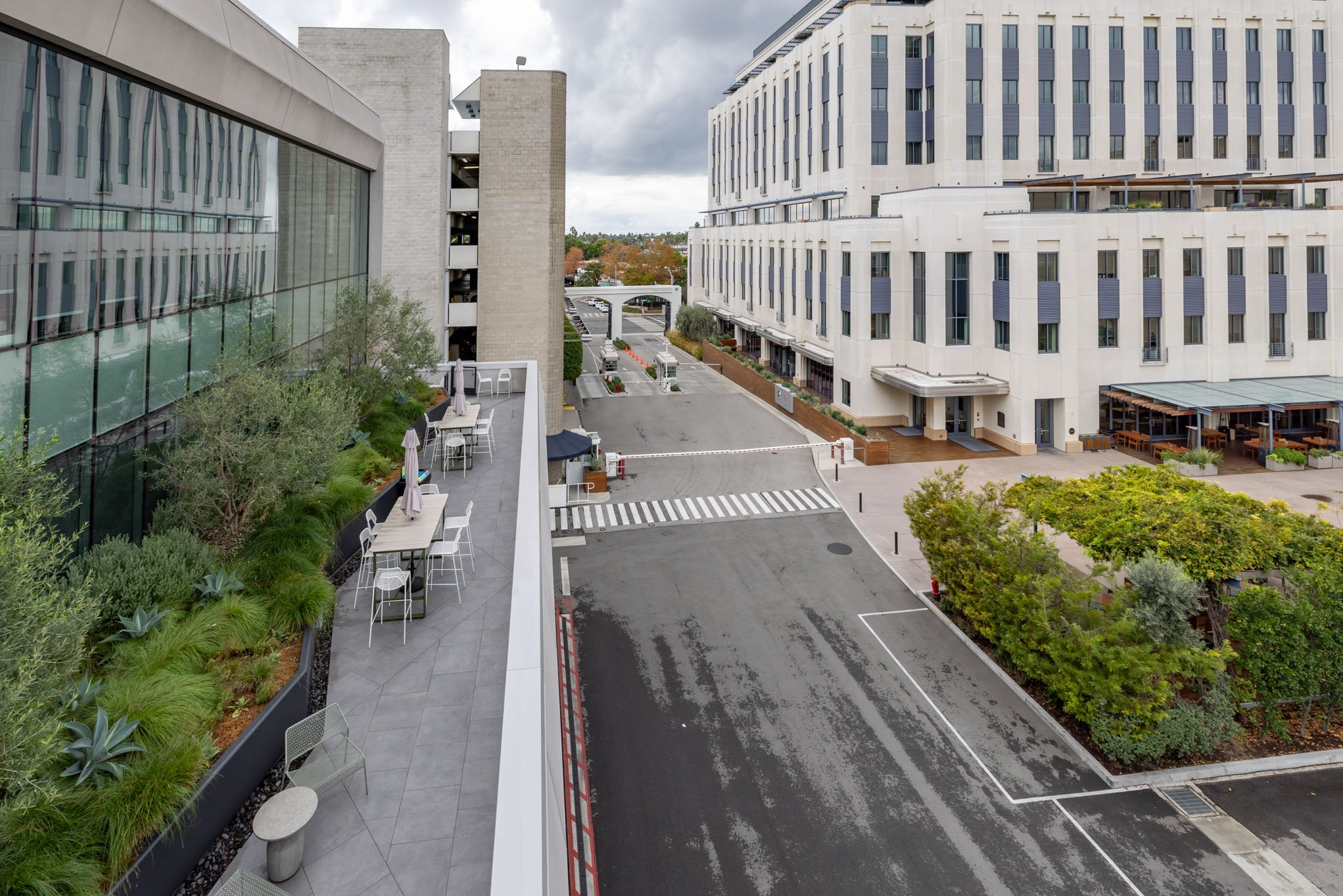 A view of an urban courtyard from an elevated position showing outdoor seating on a patio, modern buildings, greenery, and a parking area with a barrier gate.