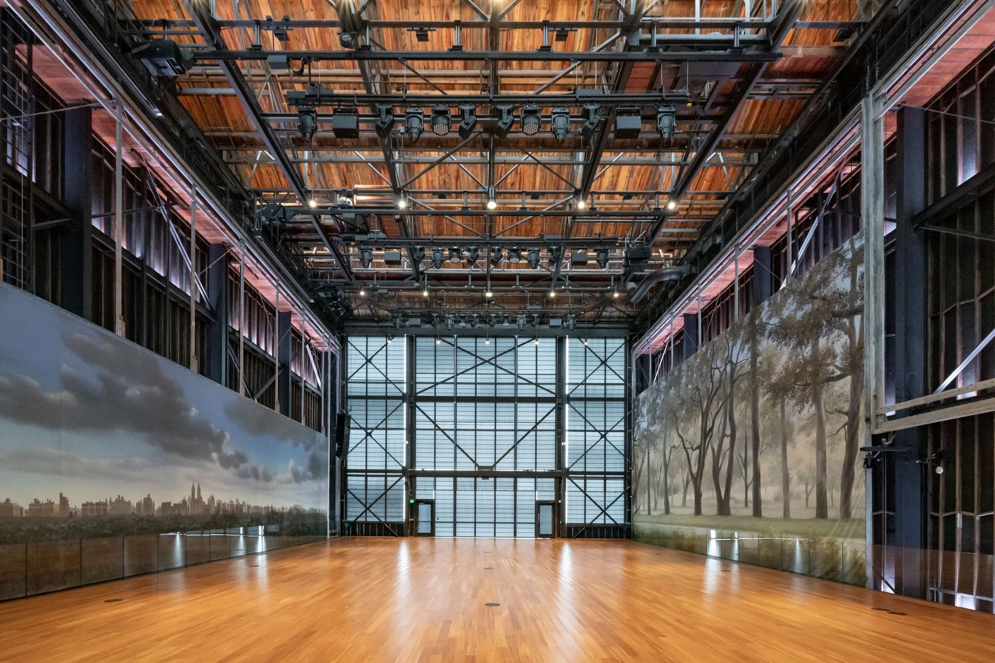 Empty indoor art gallery or exhibition hall with polished wooden floor, large murals of cityscape and forest scenery on the walls, high ceiling with exposed steel and wooden beams, and large windows at the back.