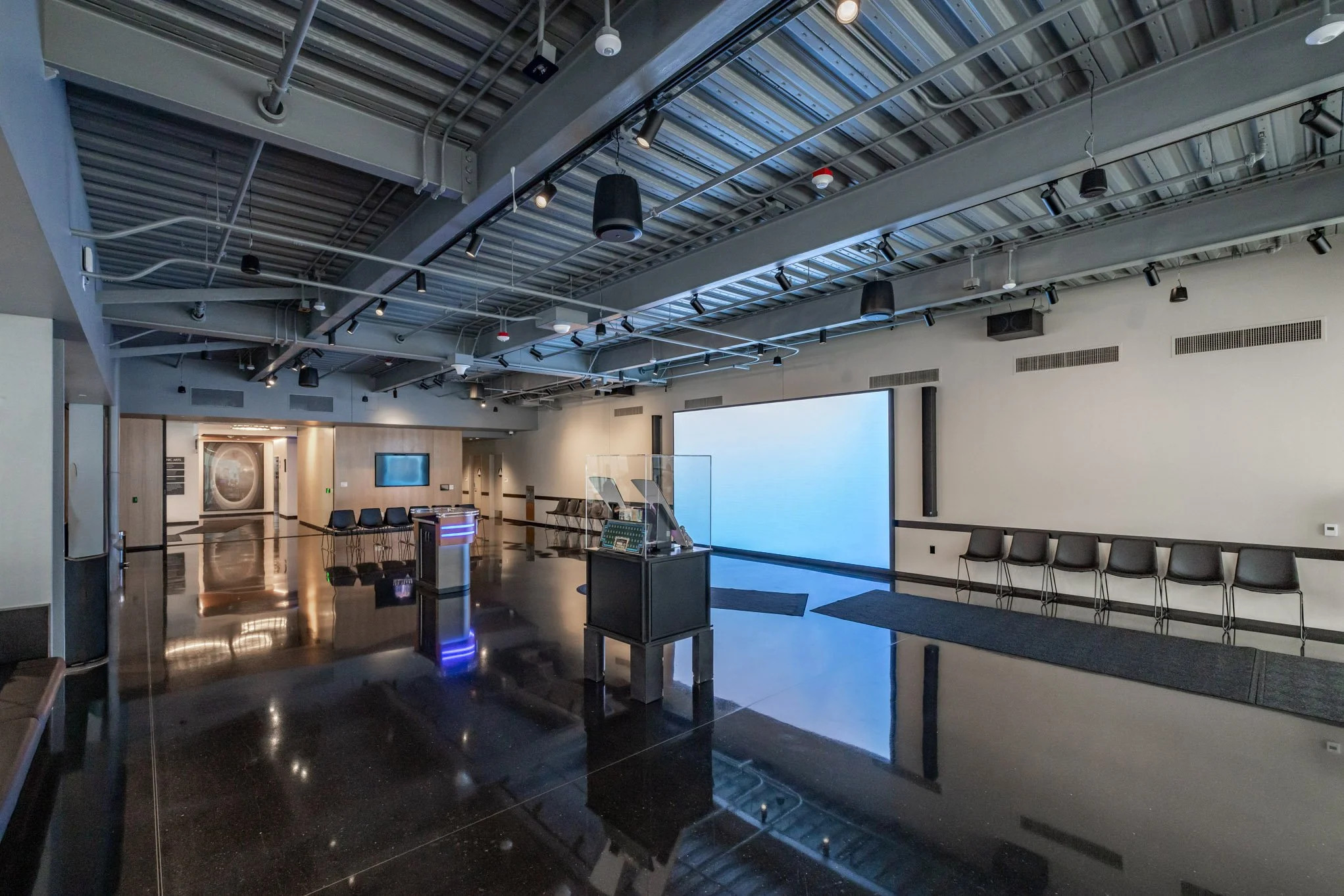 Modern, spacious conference or event room with a large blank screen, black chairs along the wall, a small table with electronic equipment, and a polished black floor reflecting the room's interior.