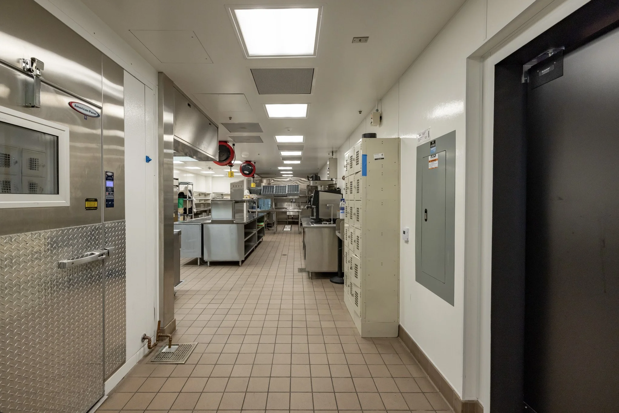 Empty commercial kitchen with stainless steel appliances, counters, lockers, and tiles on the floor.