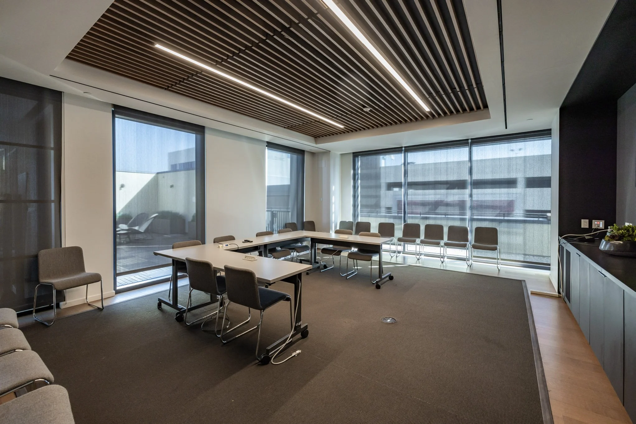 A modern conference room with multiple chairs around white tables, large windows with blinds, a wood-paneled ceiling, and a brown carpeted floor.