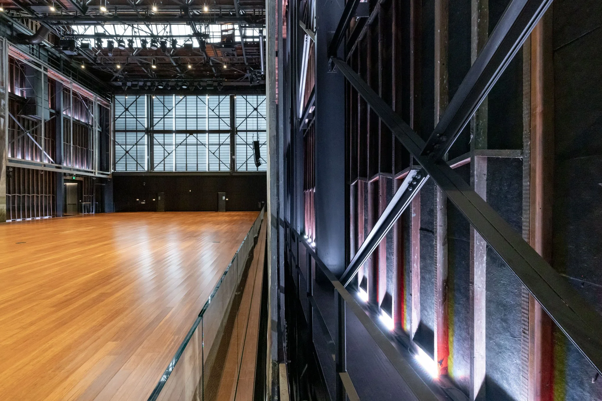 Empty indoor stage with wooden floor, large windows, and metal structural elements.