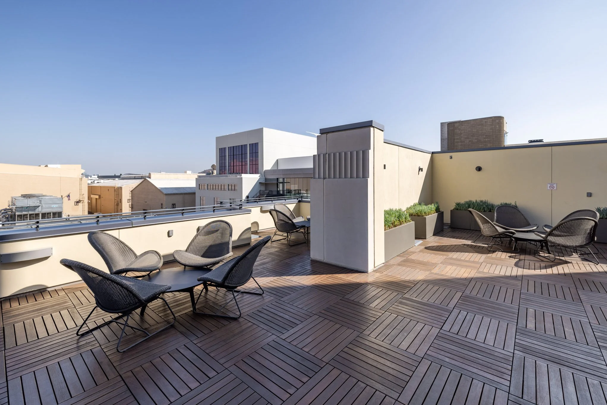 Rooftop terrace with wooden flooring, outdoor chairs and tables, potted plants, and city buildings in the background under a clear blue sky.