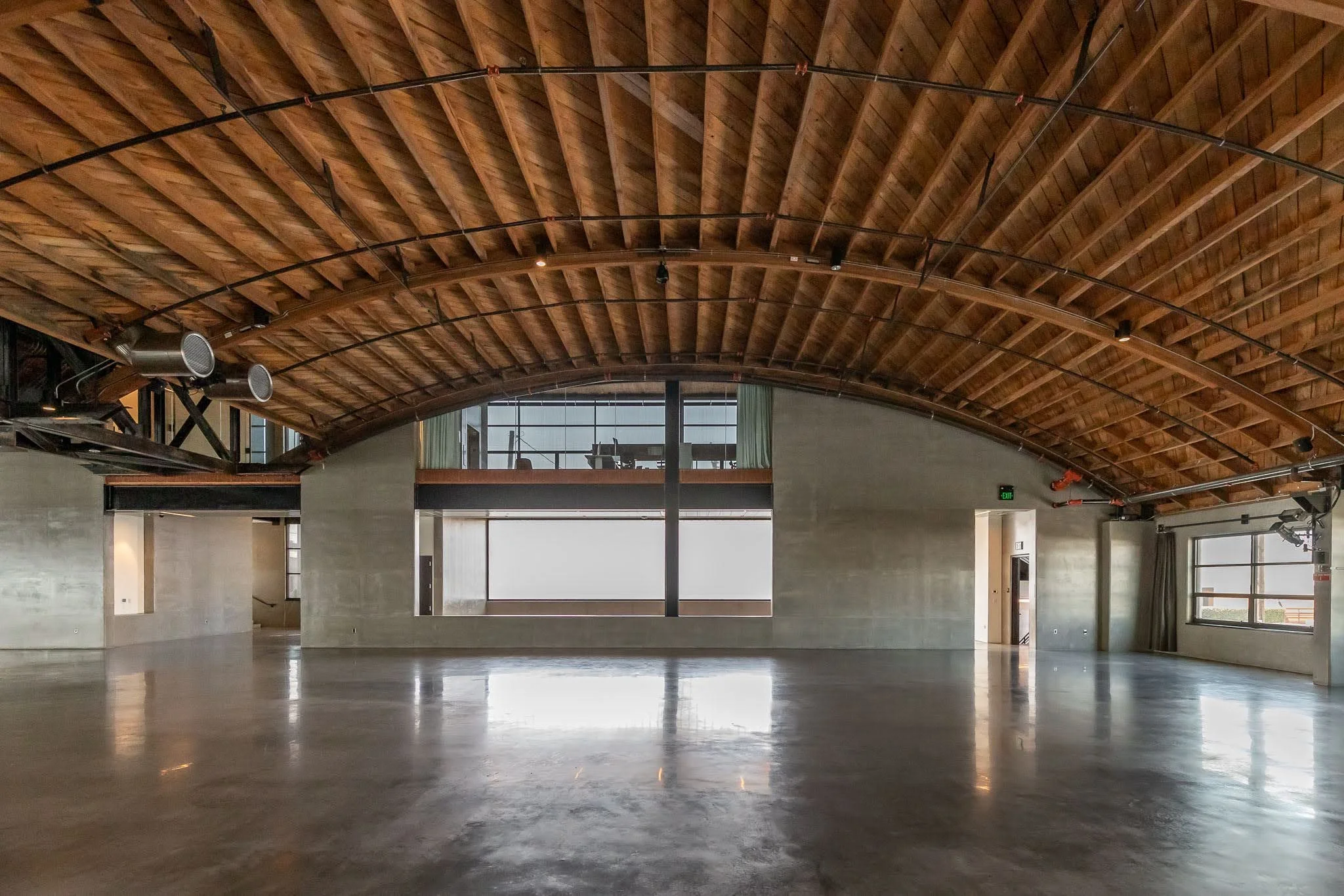 Interior view of an empty industrial-style building with a high wooden arched ceiling, large windows, concrete walls, and a polished concrete floor.