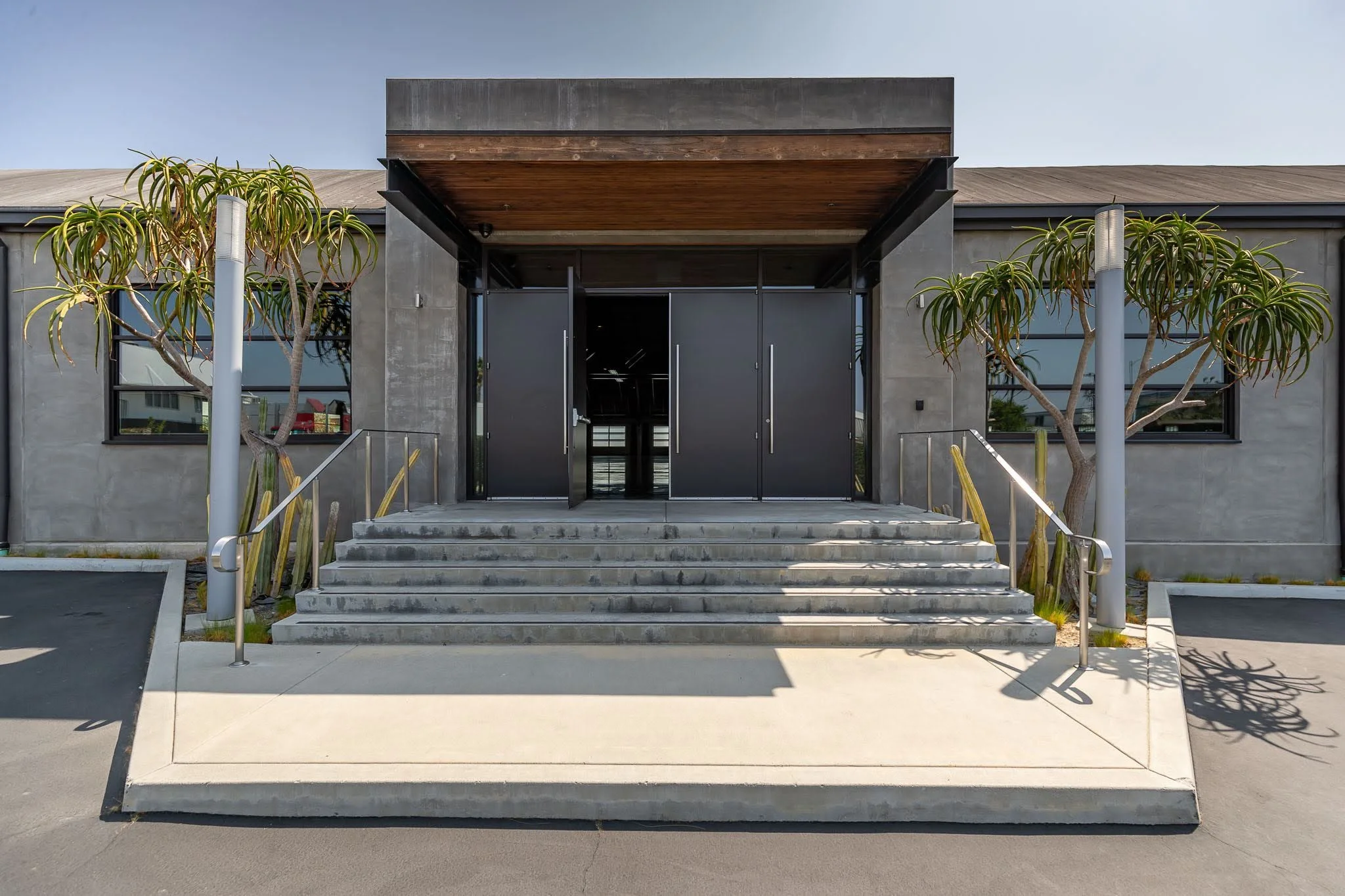Modern building entrance with concrete steps, black doors, large windows, and surrounding plants.