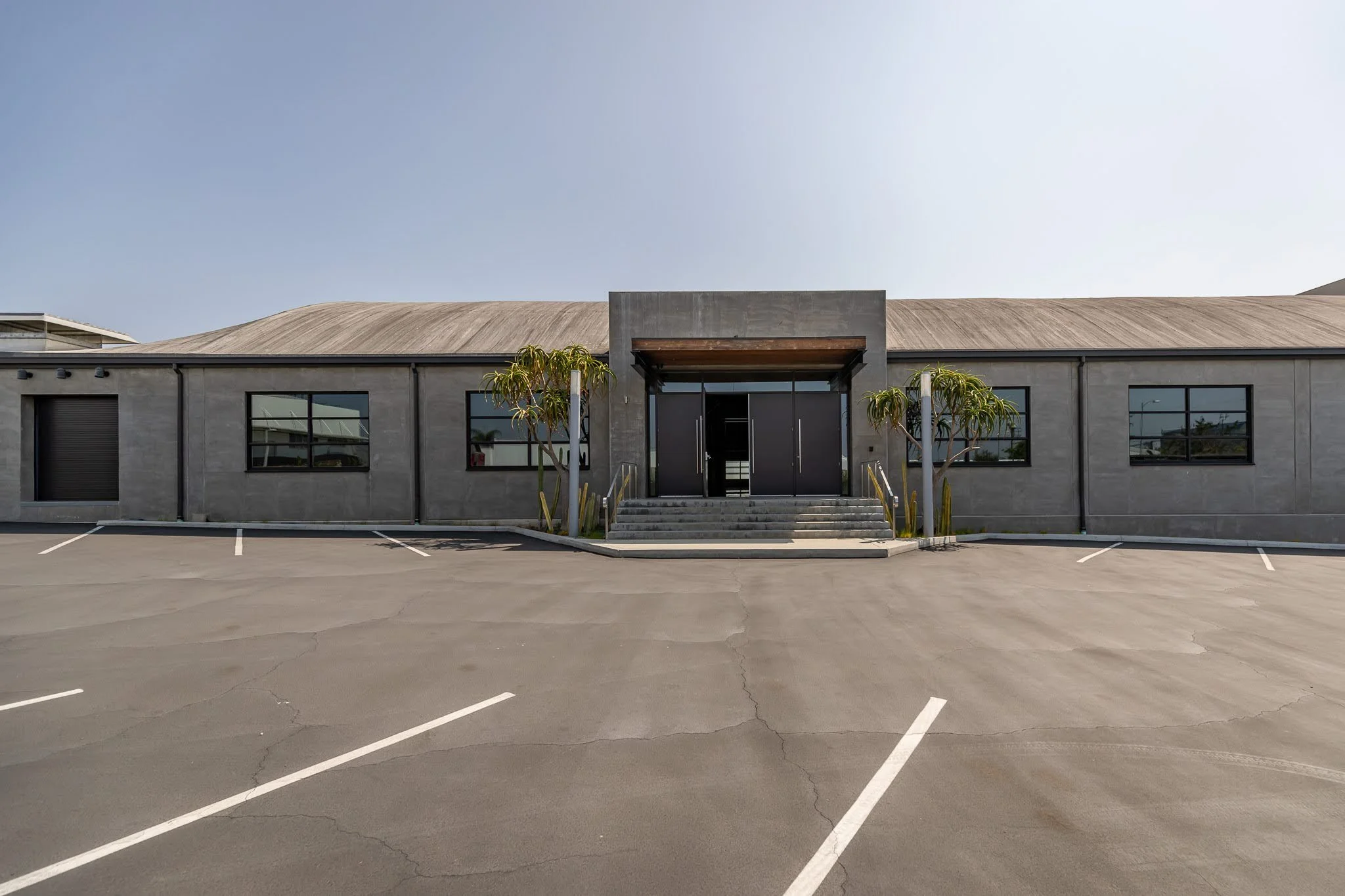 Empty parking lot in front of a modern gray building with steps and glass double doors, two small trees flanking the entrance, and clear sky overhead.