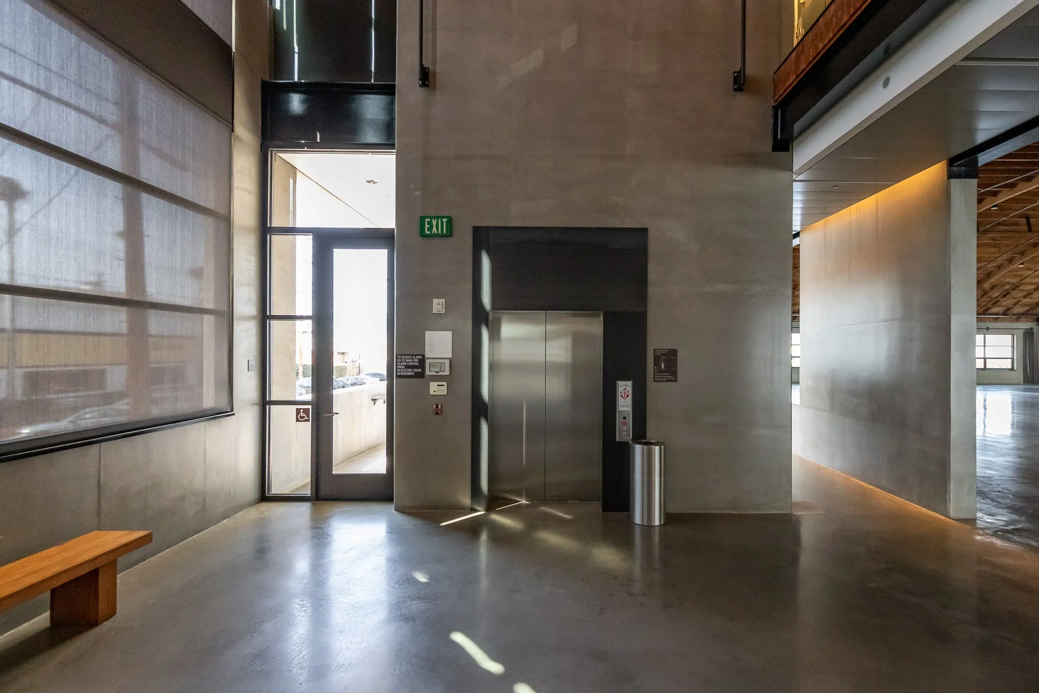 Modern indoor space with an elevator, door, exit sign, window, and bench, featuring concrete floors and walls with natural light.