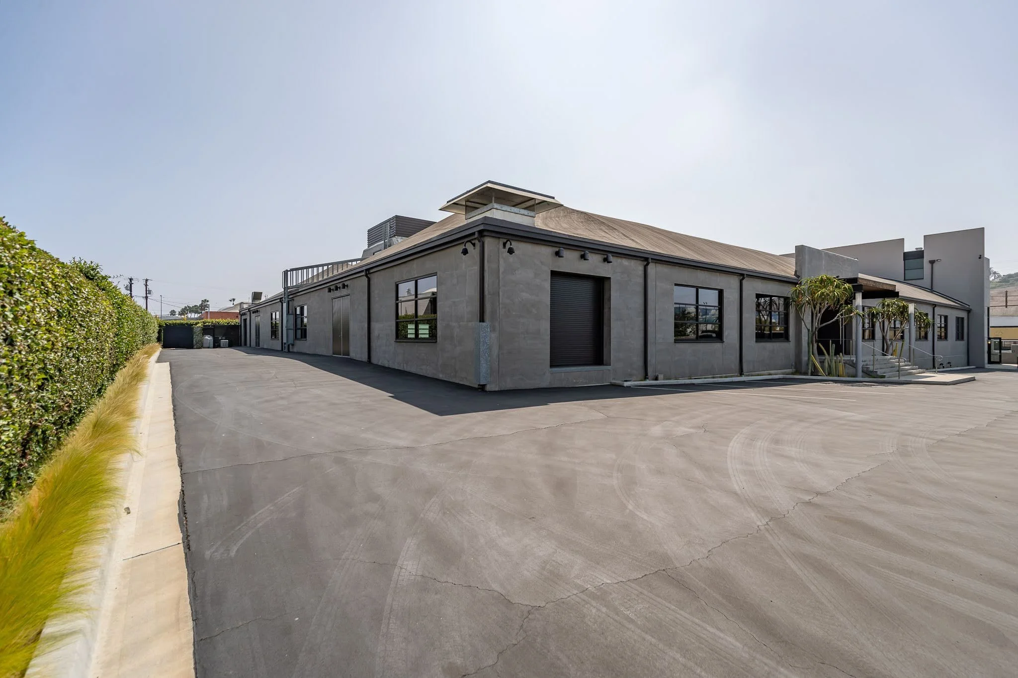 Exterior of a single-story commercial building with a large, open paved parking lot, some small palm trees near the entrance, and a hedge on the left side under a clear sky.