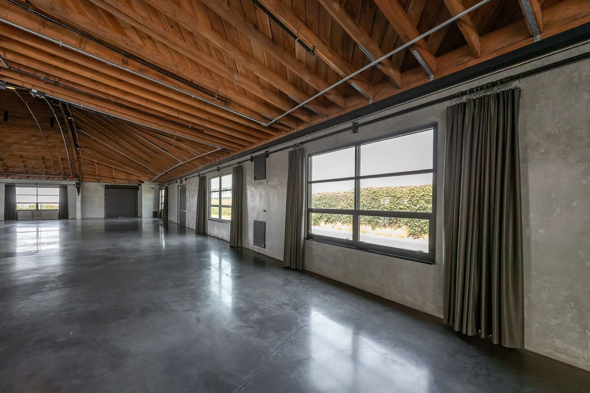 Empty warehouse or studio with polished concrete floor, large windows with curtains, and a wooden ceiling with exposed beams and metal supports.