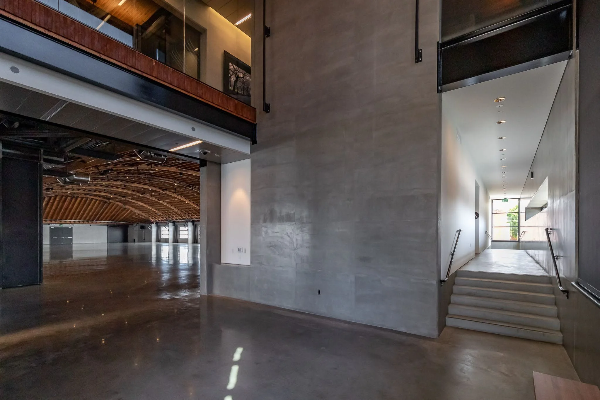 Interior of a modern building with polished concrete floors, a large open space, a staircase leading to a bright doorway, and an upper level with glass railing and wood paneling ceiling.