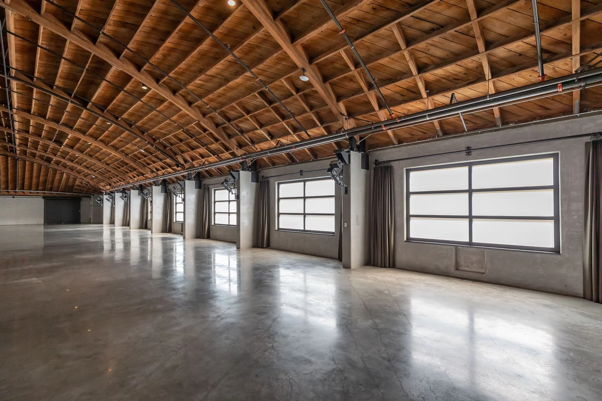 Empty industrial-style event space with concrete floors, large windows with curtains, and a wooden arched ceiling with exposed beams and lighting.