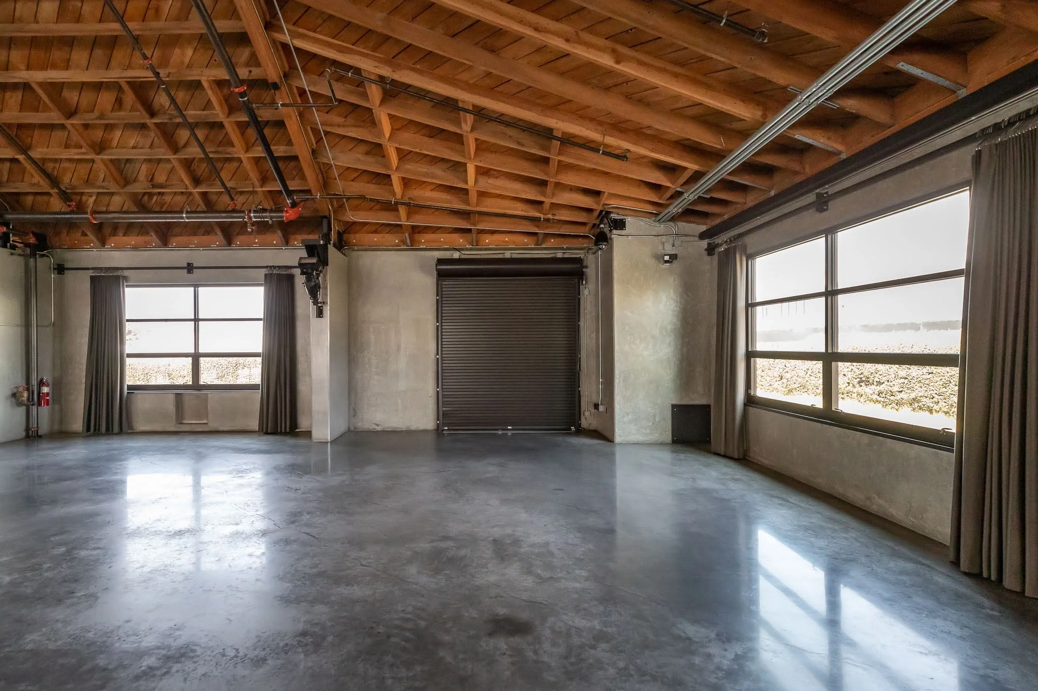 Empty industrial or commercial space with polished concrete floor, high wooden ceiling with exposed beams, large windows with curtains, and a roll-up door.