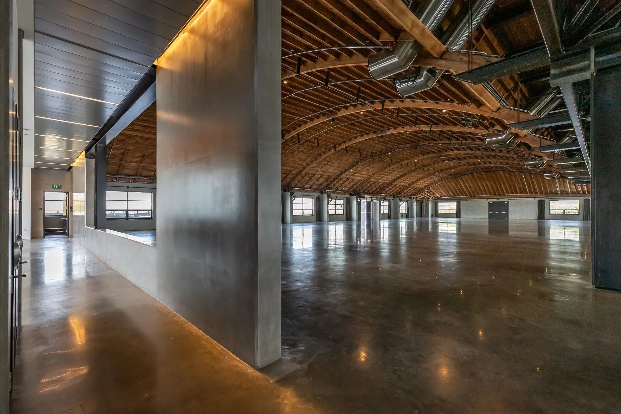 Empty indoor space with polished concrete floors, a curved wooden ceiling with exposed beams, large windows, and industrial ventilation ducts.