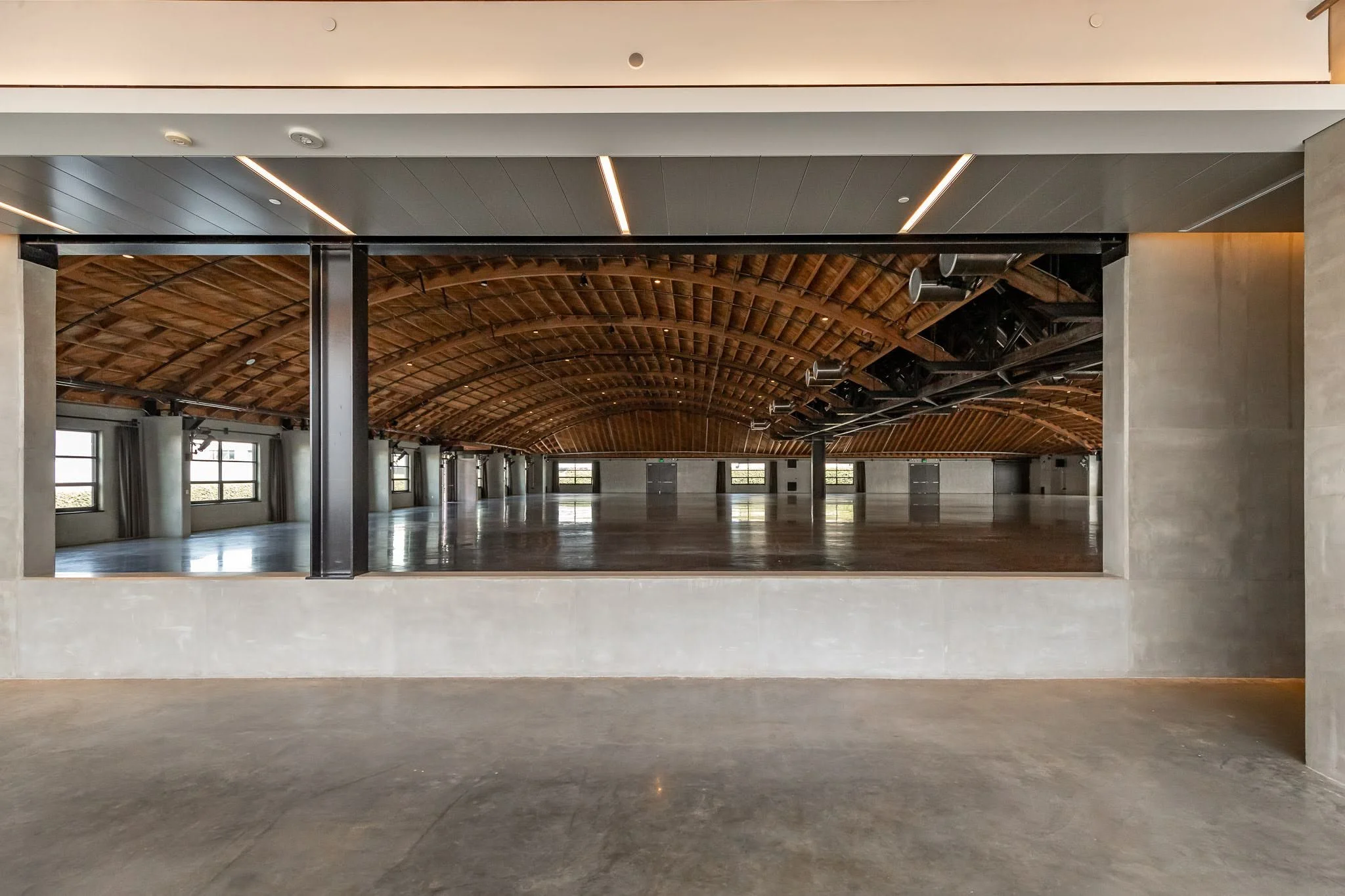 Empty large indoor space viewed through a rectangular opening with a polished concrete floor, a high arched wooden ceiling, and multiple windows along the sides.