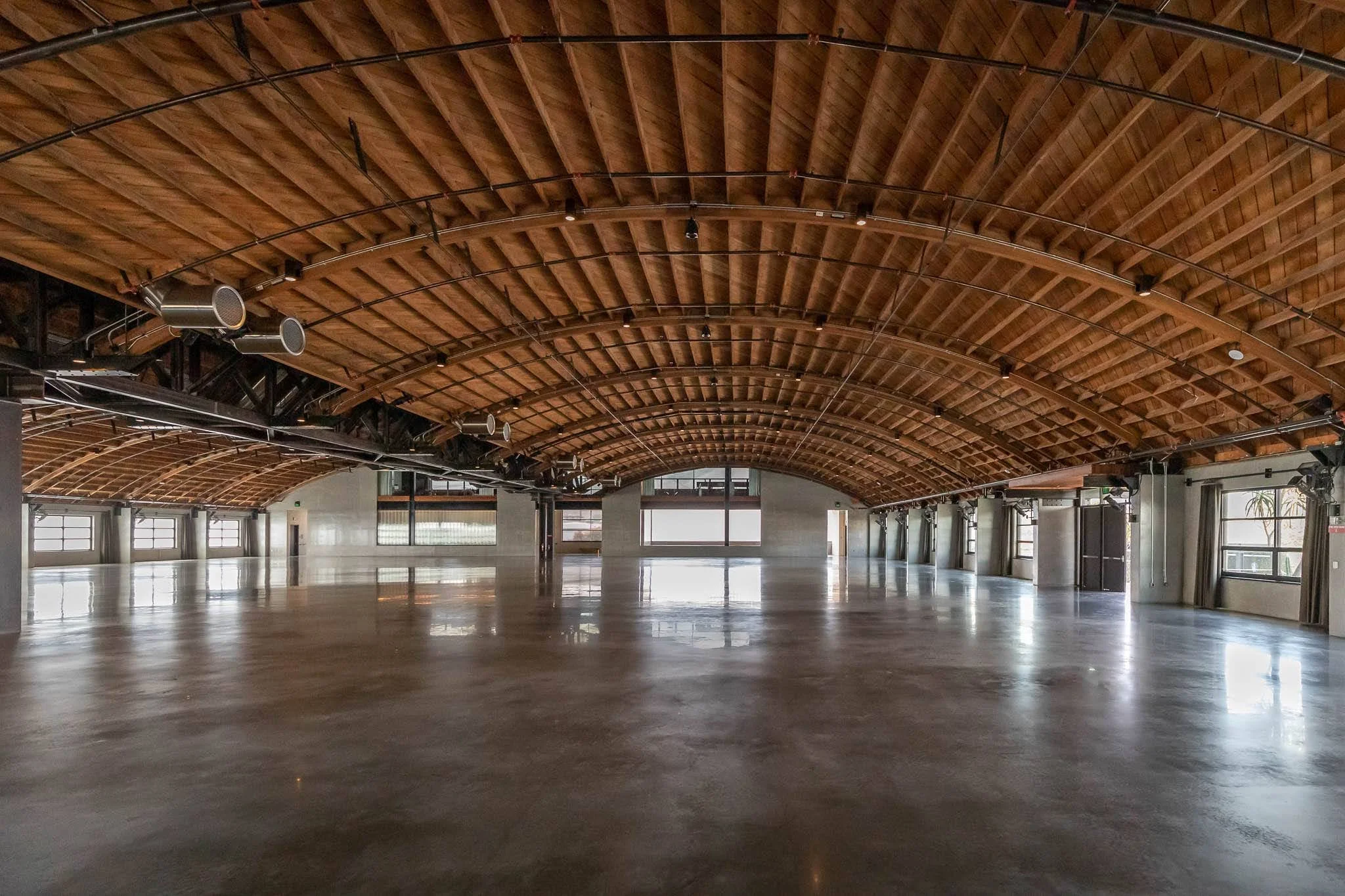 Empty large open space with polished concrete floors and a curved wooden ceiling, with multiple windows along the sides allowing natural light.