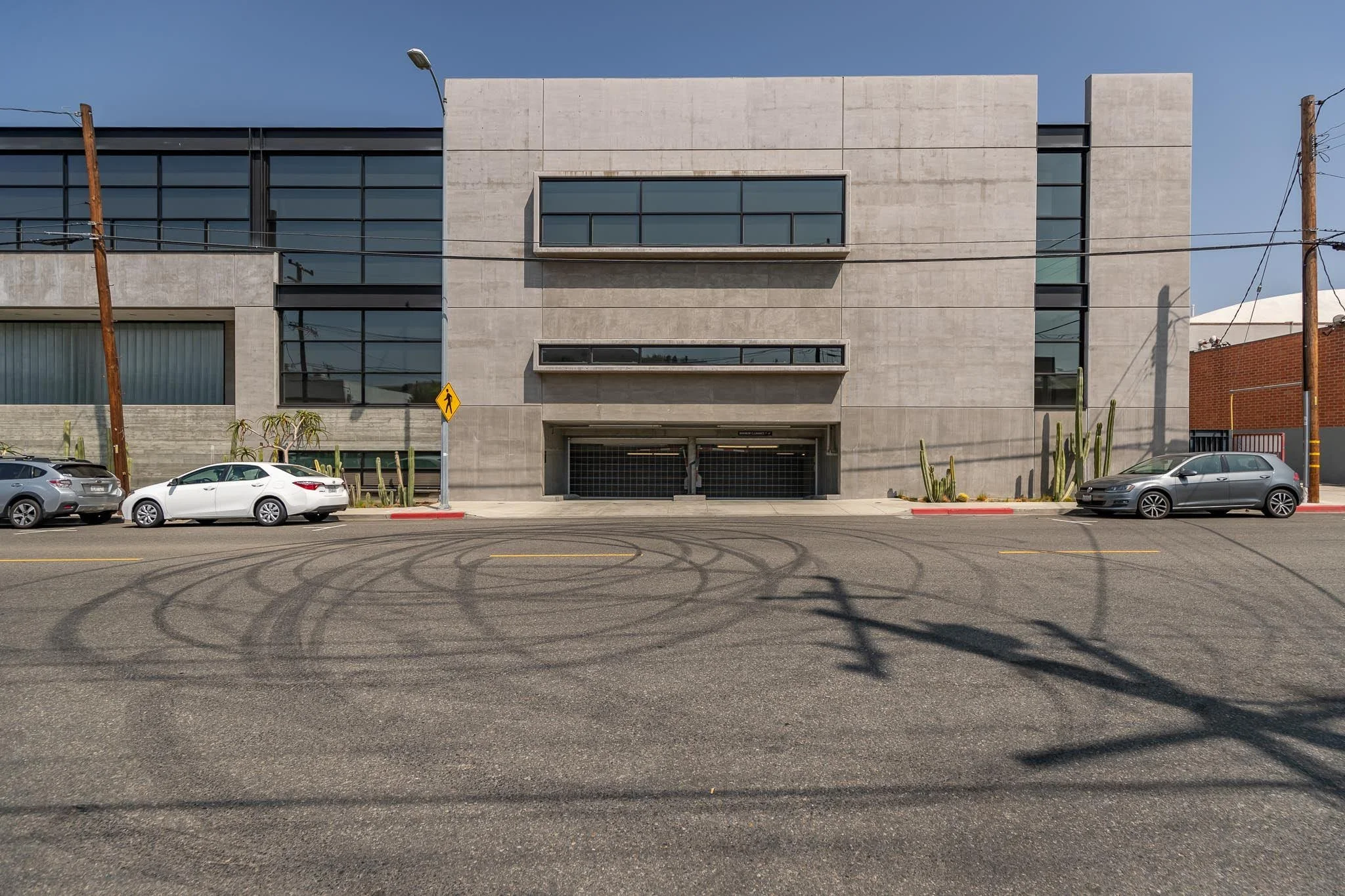 Modern gray concrete building with large glass windows and parking lot with cars, tire marks on the street, and utility poles.
