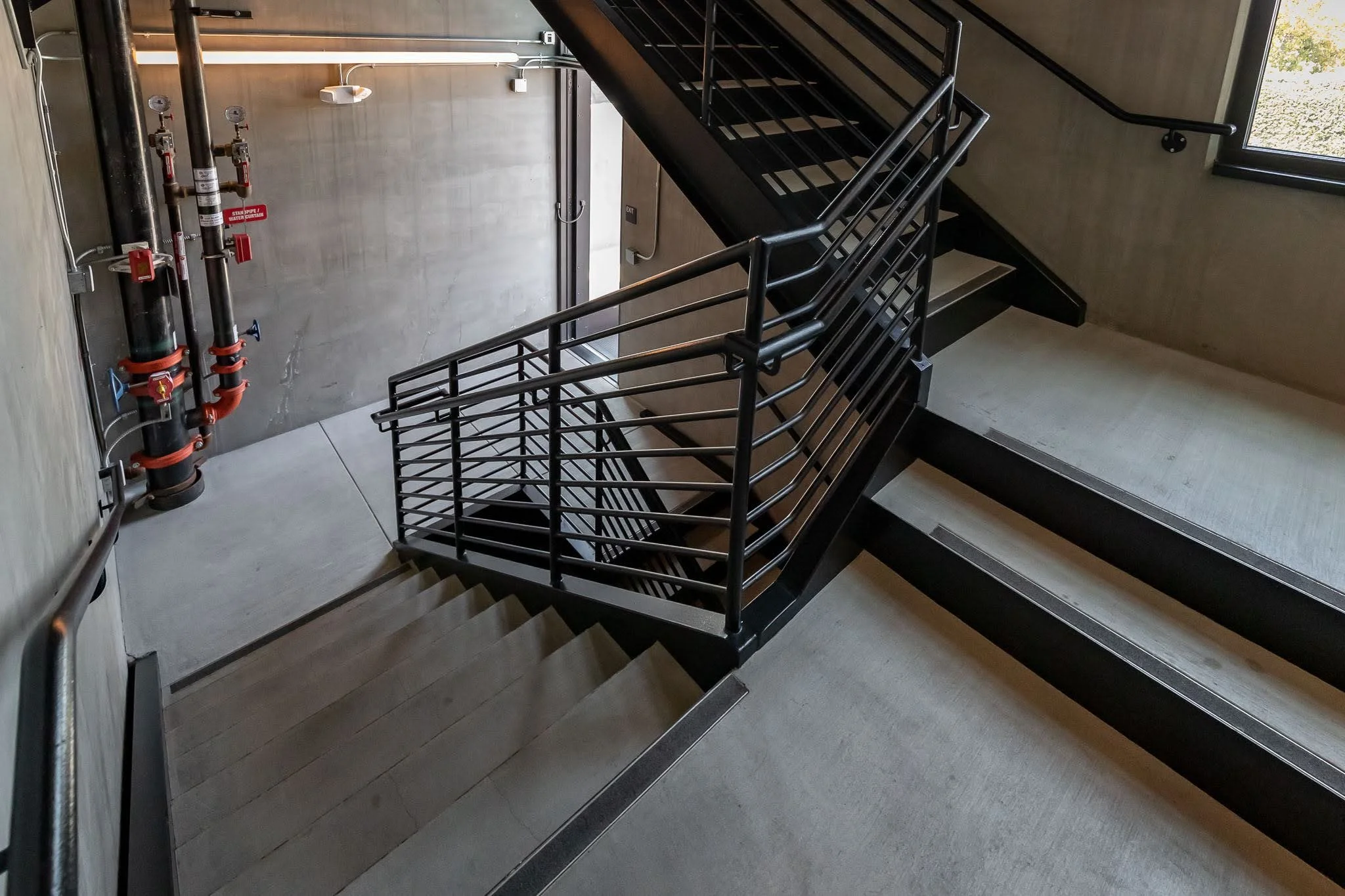 View of a staircase with black metal railings inside a building, with fire sprinkler pipes and a fire alarm on the wall nearby, and an exit door at the bottom of the staircase.