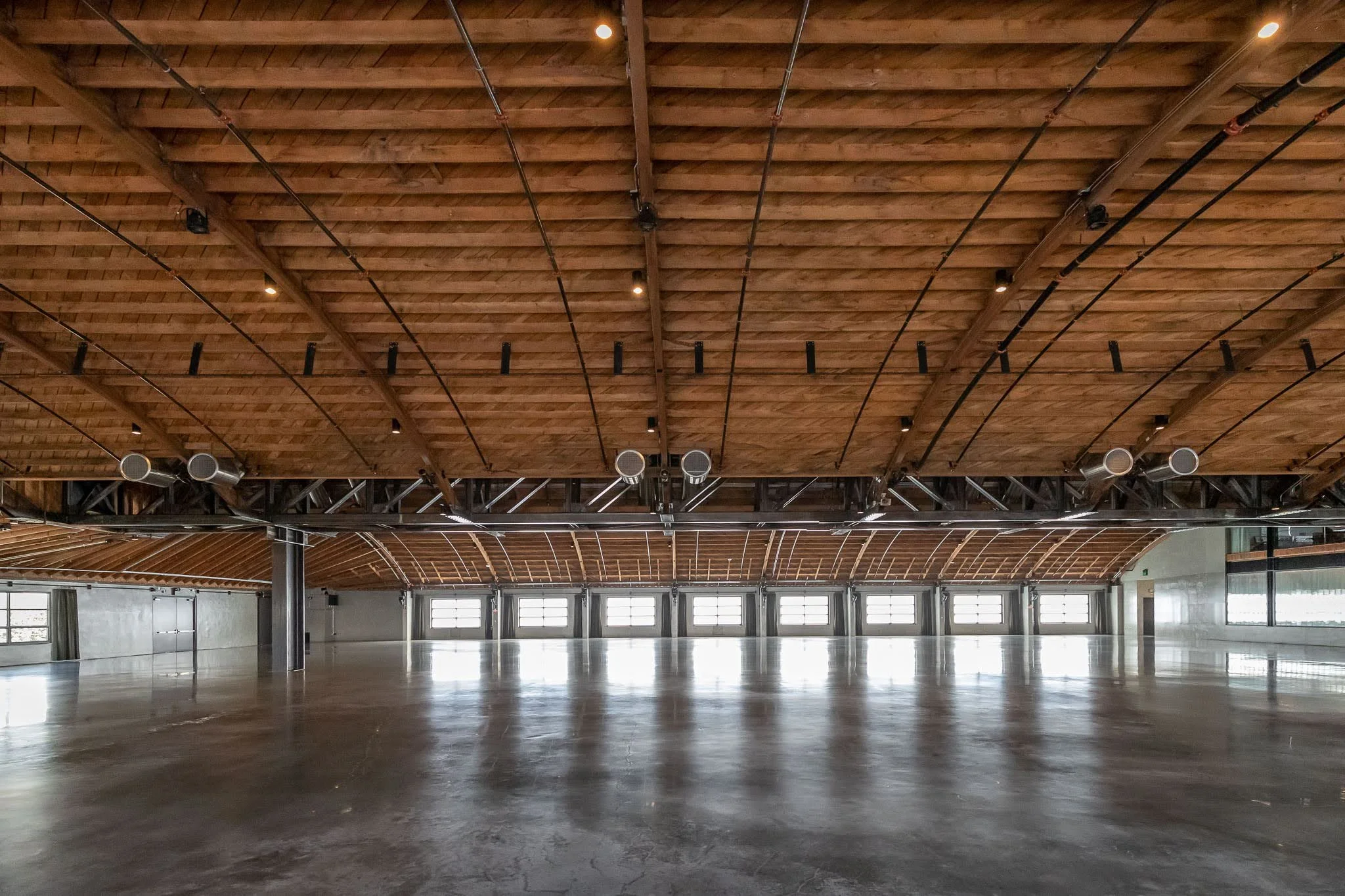 Empty large indoor space with polished concrete floor, wooden vaulted ceiling, and multiple windows along the far wall allowing natural light to enter.