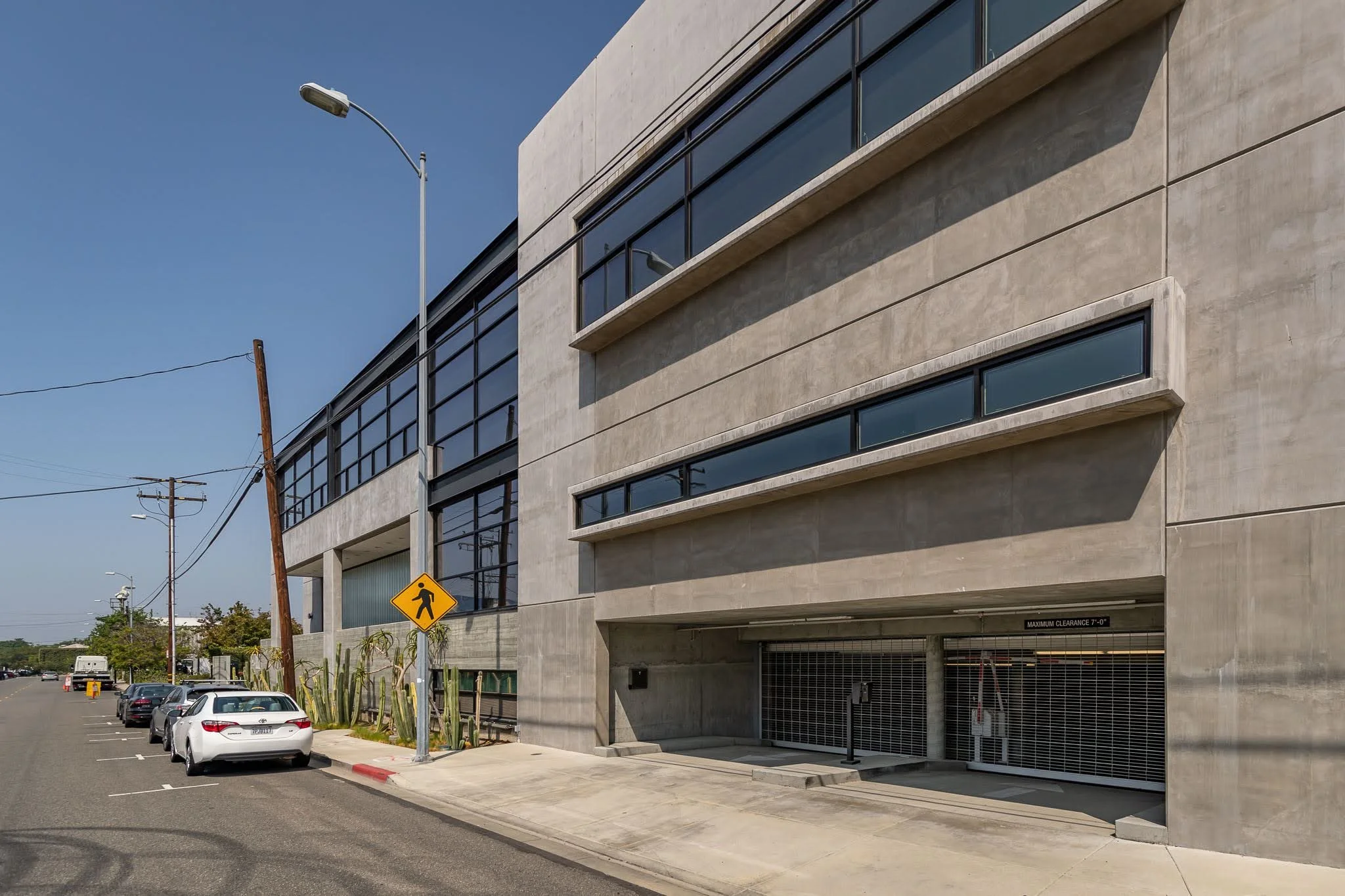 Modern multi-story concrete building with large horizontal windows, a parking garage entrance, parked cars, and a pedestrian crossing sign on a sunny day with a clear blue sky.