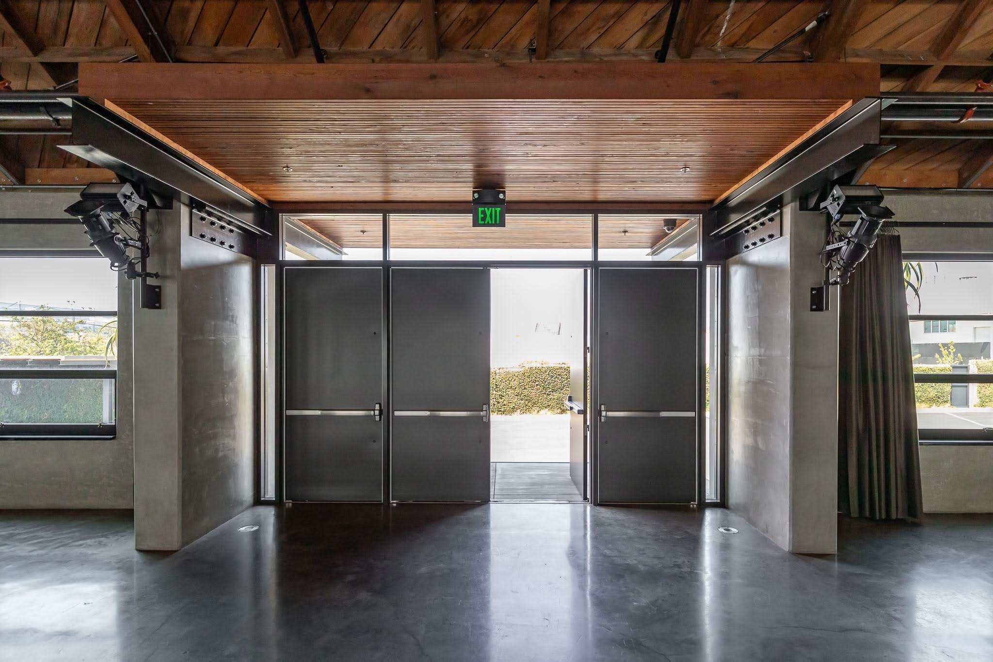 Interior of a modern building with double glass doors, an exit sign above, and windows on each side, revealing an outdoor area with trees and shrubs.