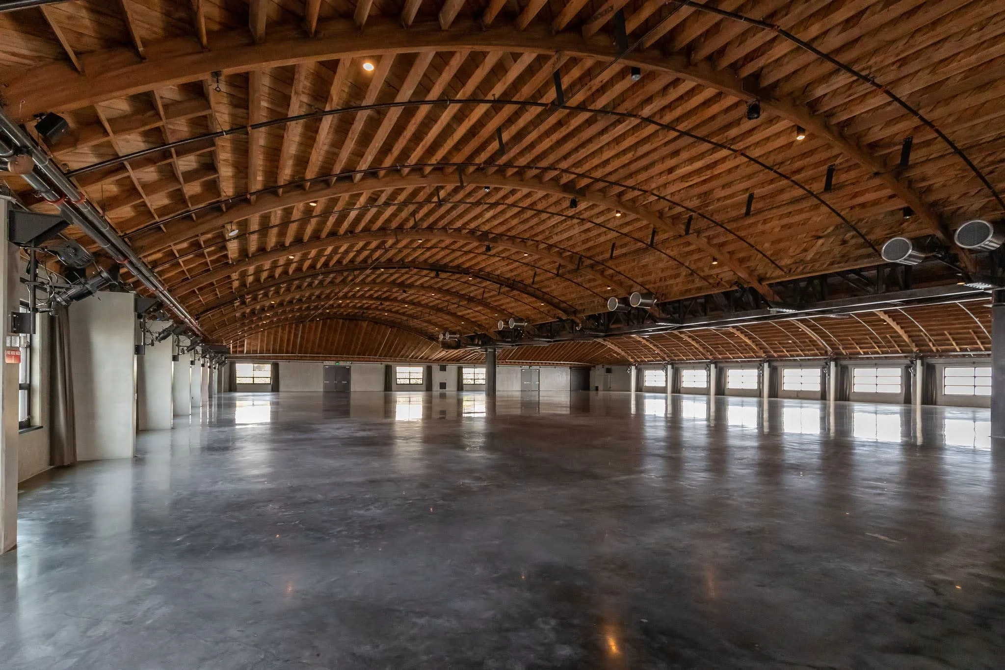 Large, empty event hall with polished concrete floor, wooden arched ceiling, and multiple windows along the walls, with stage lighting and ventilation systems overhead.
