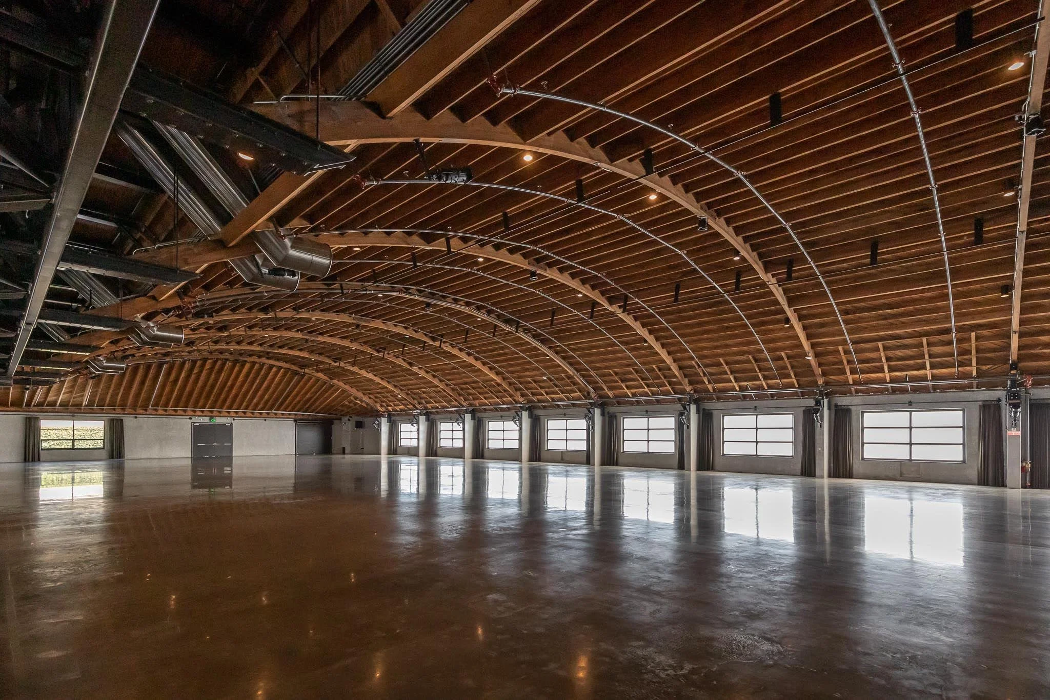 Empty indoor event space with a polished concrete floor, high wooden vaulted ceiling with exposed beams and HVAC ducts, and large windows with gray curtains.