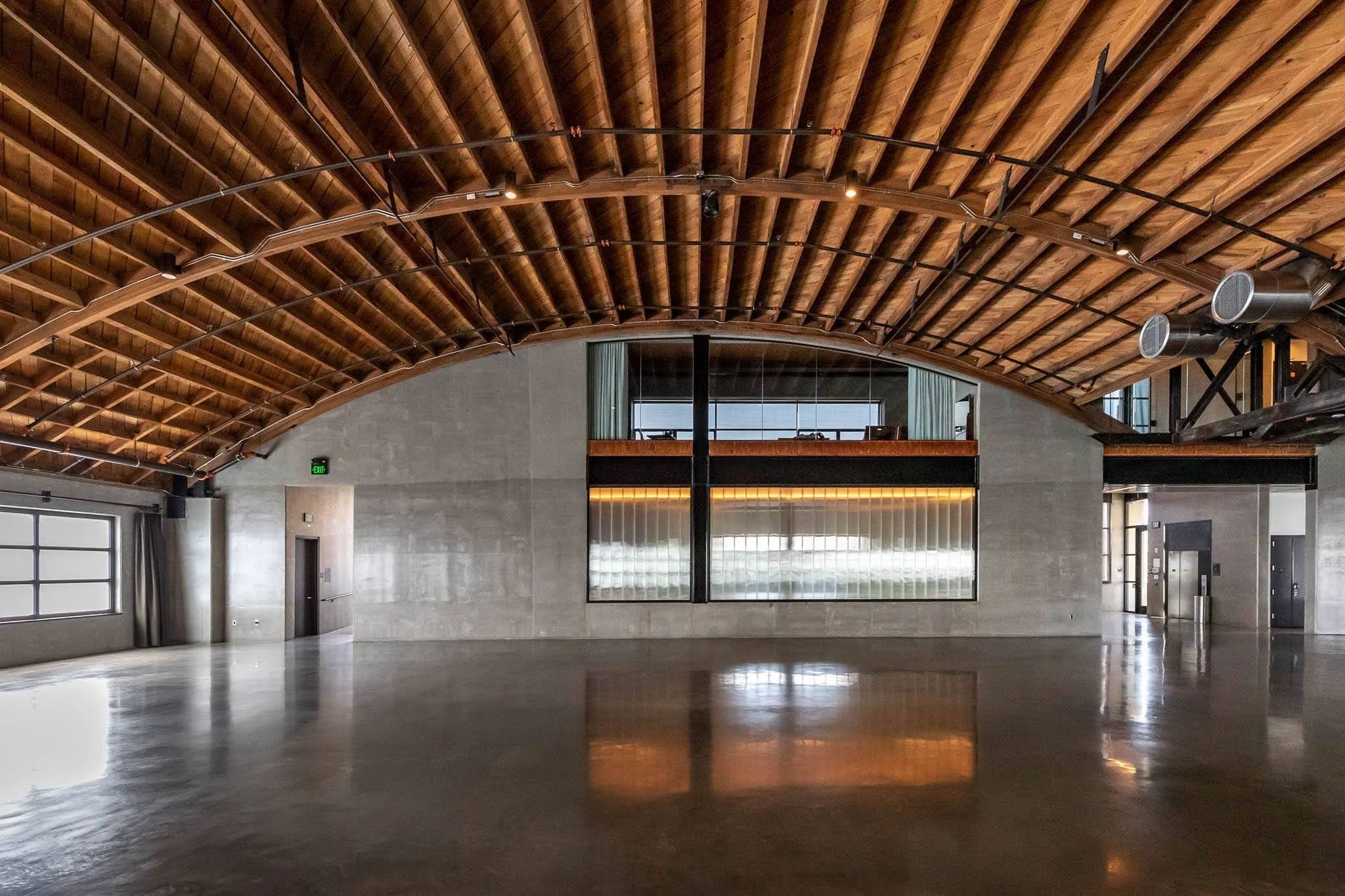 Empty large room with polished concrete floor, wooden arched ceiling, large windowed wall, elevator doors, and industrial lighting.