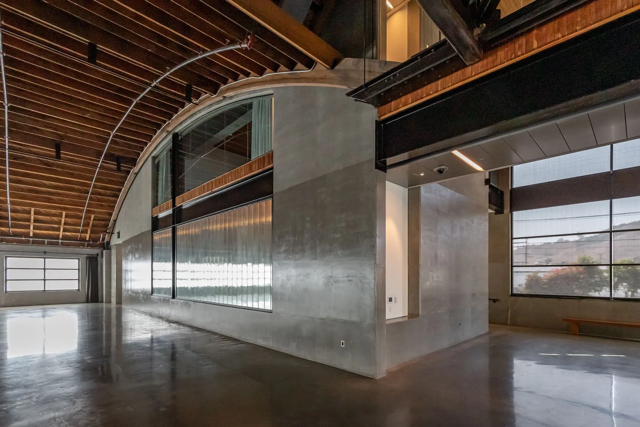 Interior of a modern building with a high wooden ceiling, large windows with blinds, and a curved concrete wall.