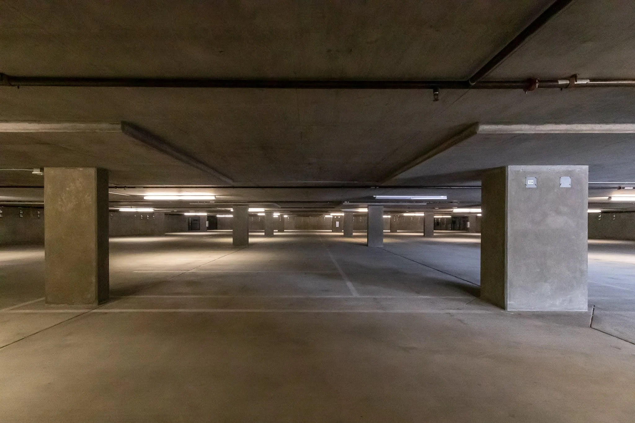 Empty underground parking garage with concrete pillars and ceiling, dim lighting, and parking spaces marked on the floor.