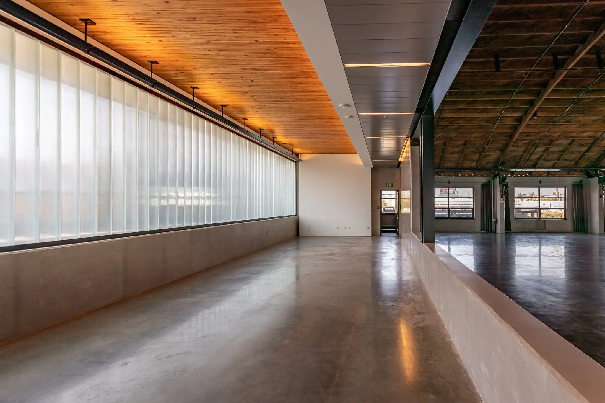 Empty interior space with a polished concrete floor, large frosted window, and wooden ceiling on one side, with a darker wooden arched ceiling on the other side, and large windows at the far end.