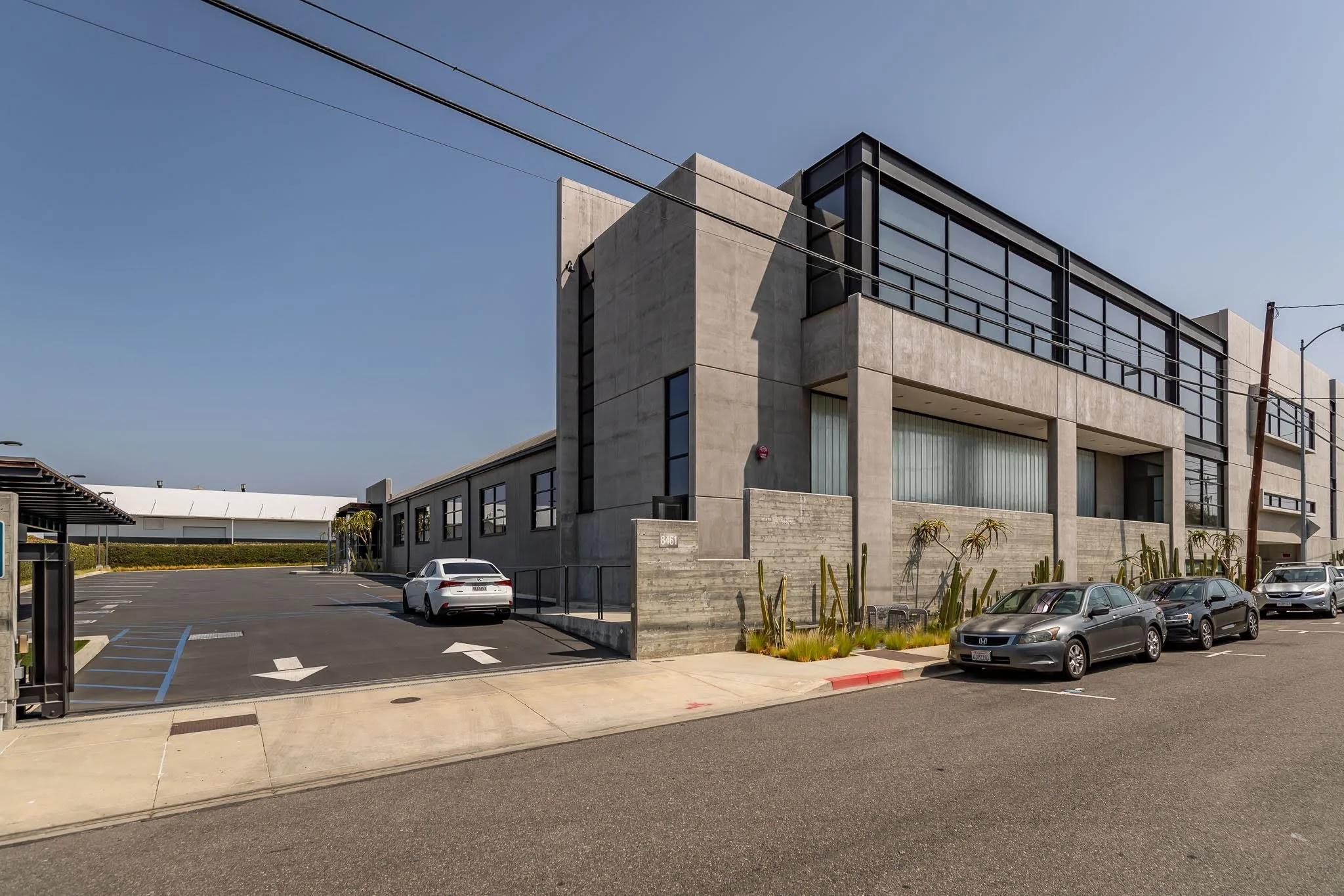 A modern concrete building with large glass windows, adjacent parking lot with several parked cars, and desert plants in front.