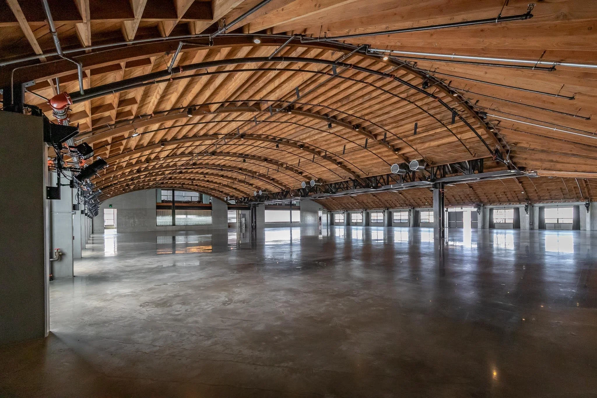 Empty spacious indoor area with polished concrete floor, large windows at the back, and an arched wooden ceiling with exposed beams and electrical conduits.