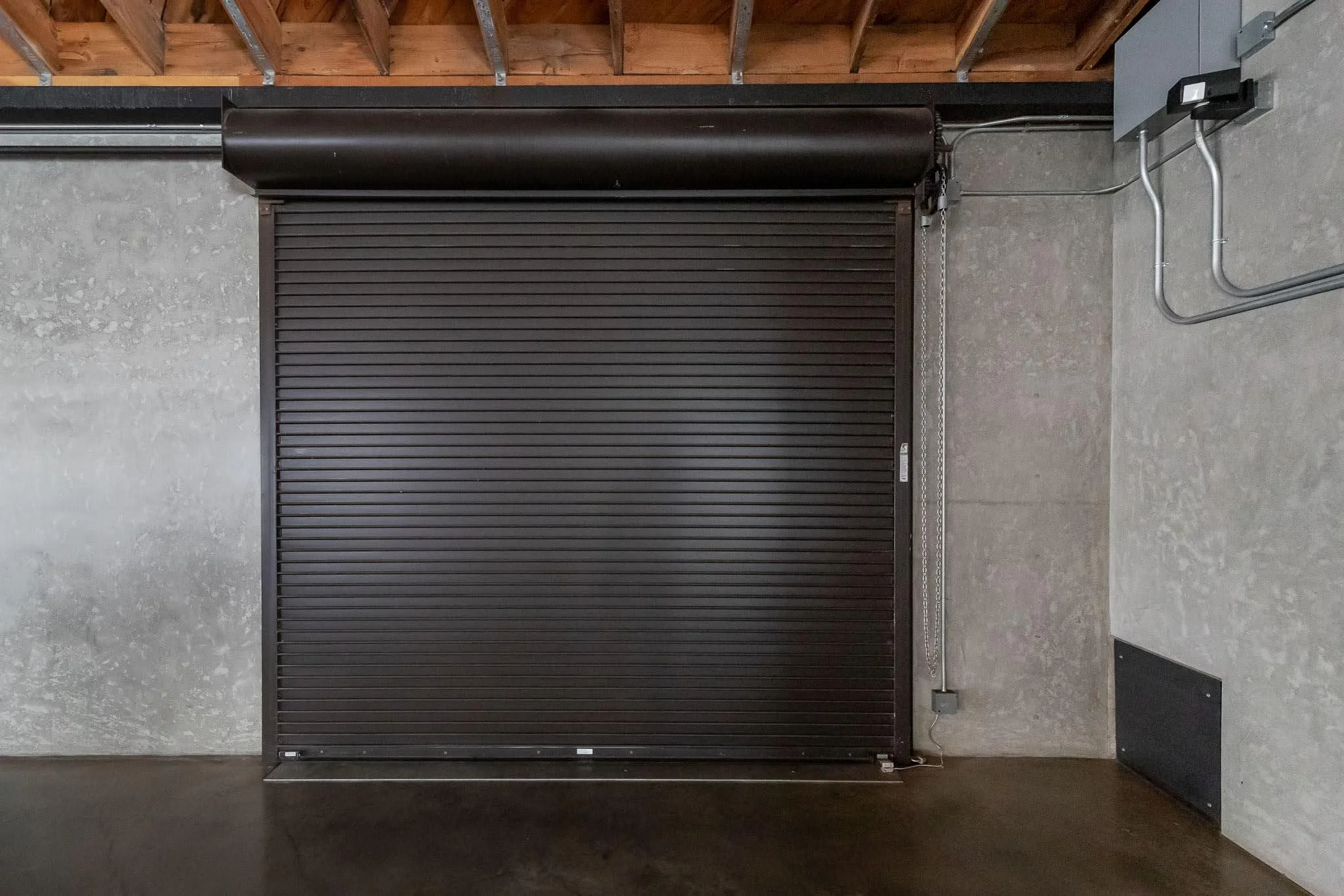 Black metal roll-up garage door closed in a concrete-walled garage with exposed electrical conduit and a ceiling with wooden beams.