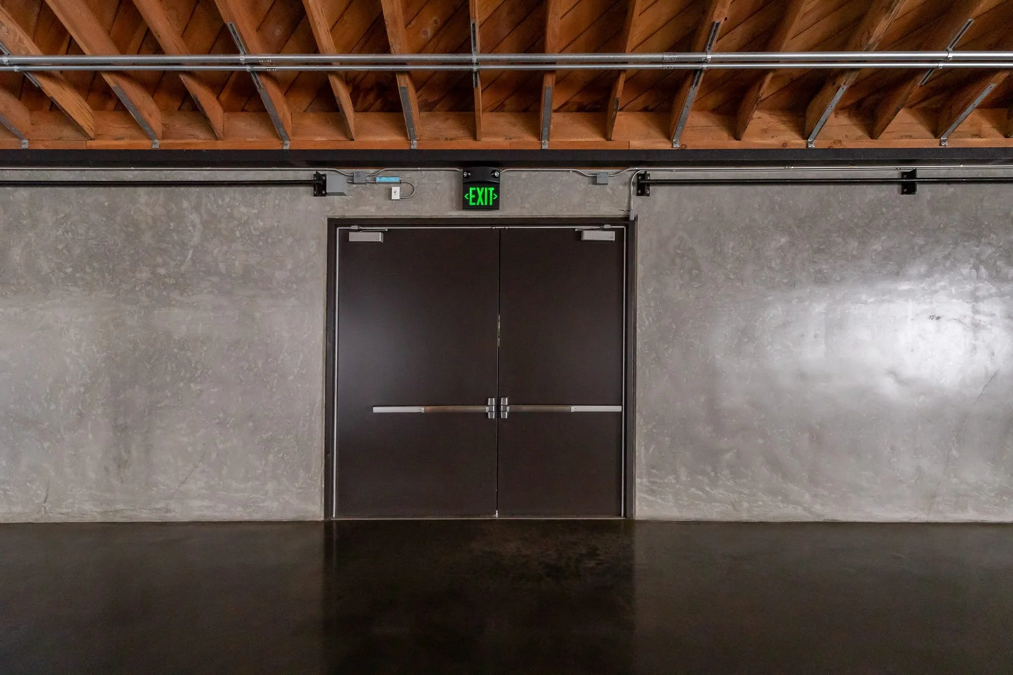 Double black door with a metal push bar, set in a concrete wall, with an illuminated green exit sign above indicating an emergency exit.