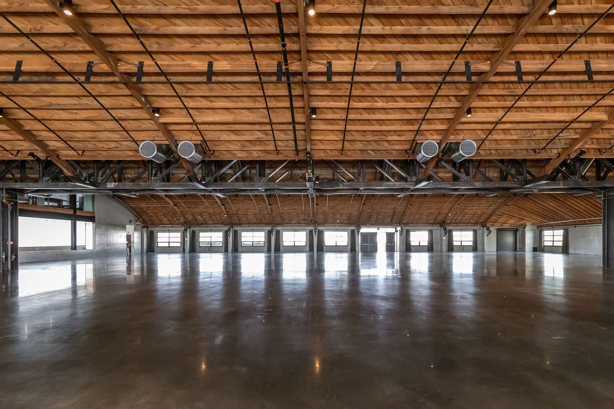 Interior of a spacious, empty event hall or warehouse with polished concrete floor, wooden ceiling with exposed beams, and large windows letting in natural light.