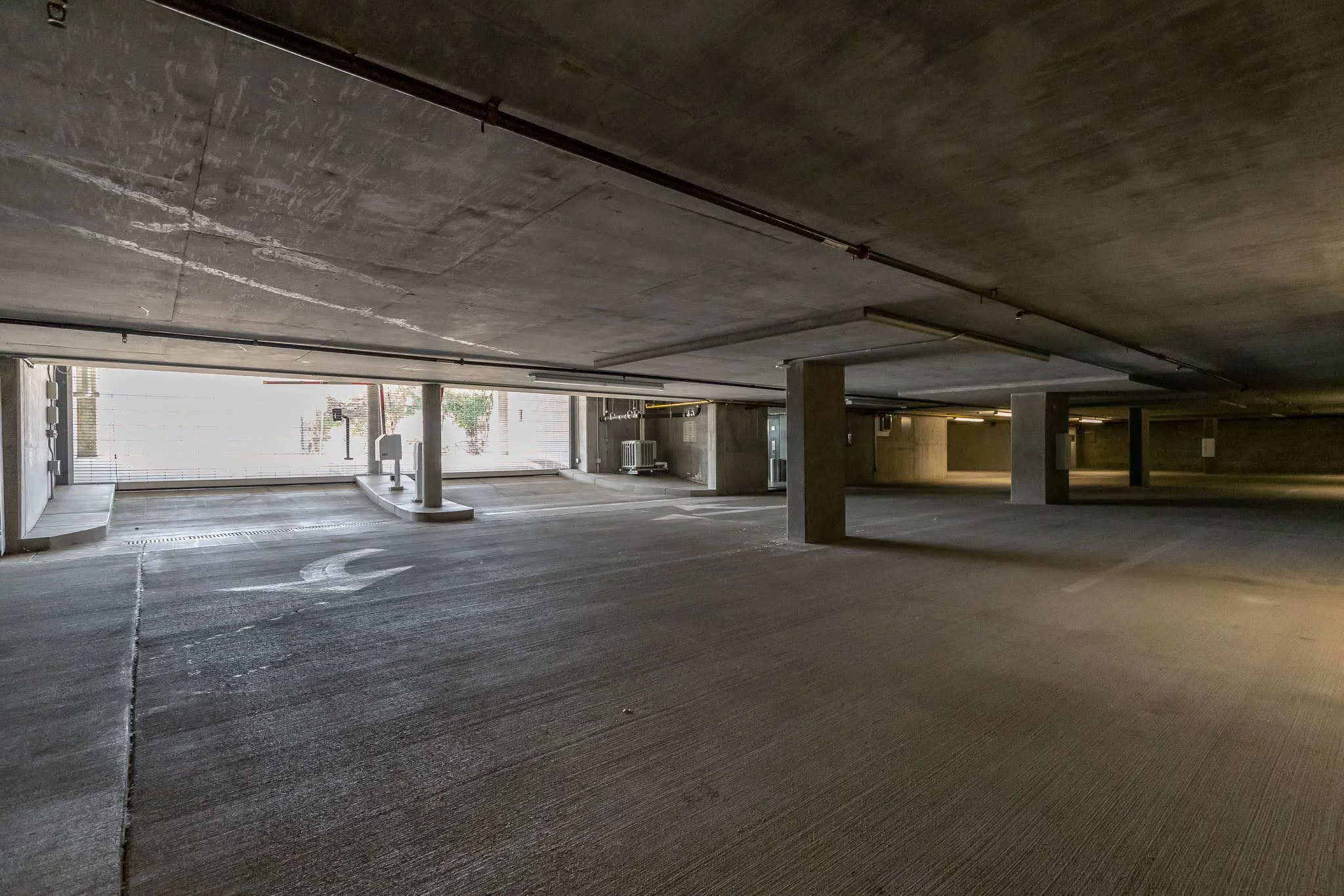 Empty indoor parking garage with concrete pillars, ceiling, and floor, illuminated by fluorescent lights with some natural light coming through an open entrance.